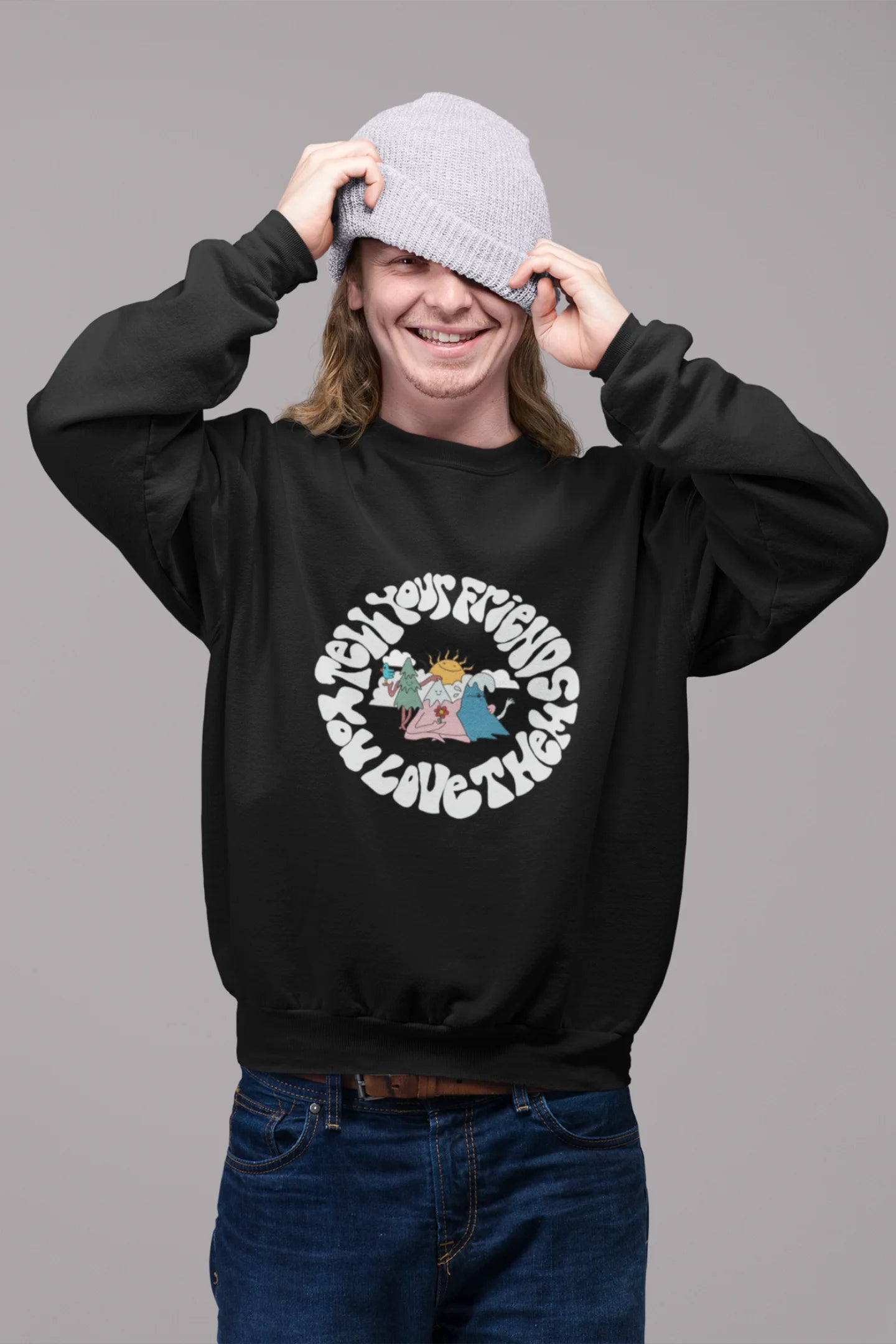 First front view of a male model wearing a black sweatshirt with a design of smiling mountains and a sunrise, and the text "Tell Your Friends You Love Them."