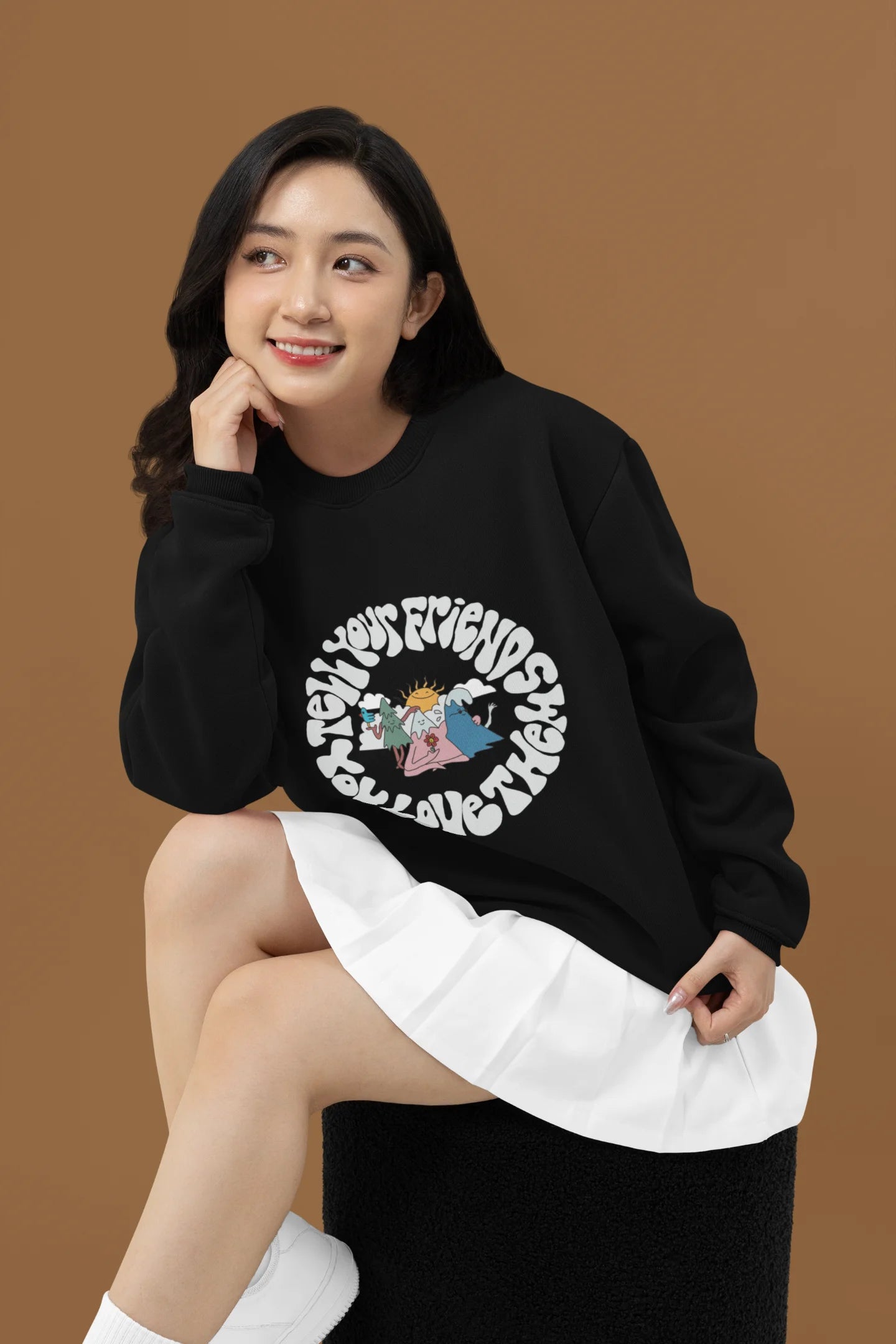 2nd front view of a female model wearing a black sweatshirt with a design of smiling mountains and a sunrise, and the text "Tell Your Friends You Love Them."