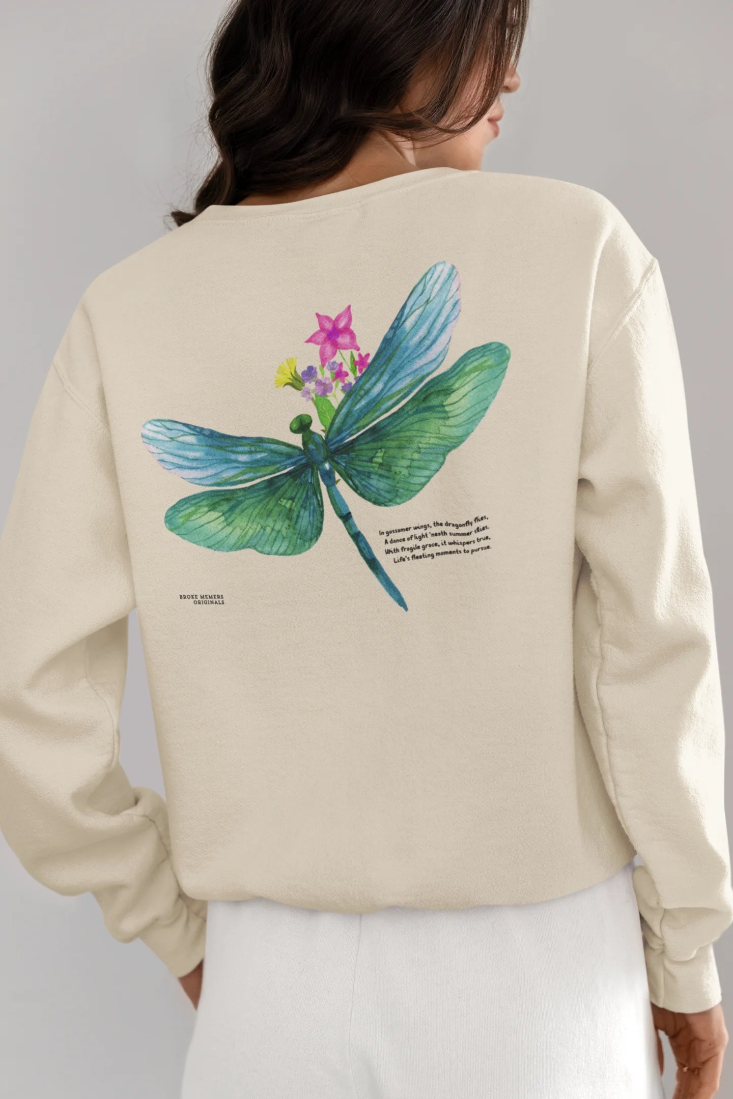 Back view of a female model wearing a cream sweatshirt featuring a nature-inspired design with a blue dragonfly perched on flowers amidst green grass. Ideal for those who love springtime aesthetics and dragonfly imagery.