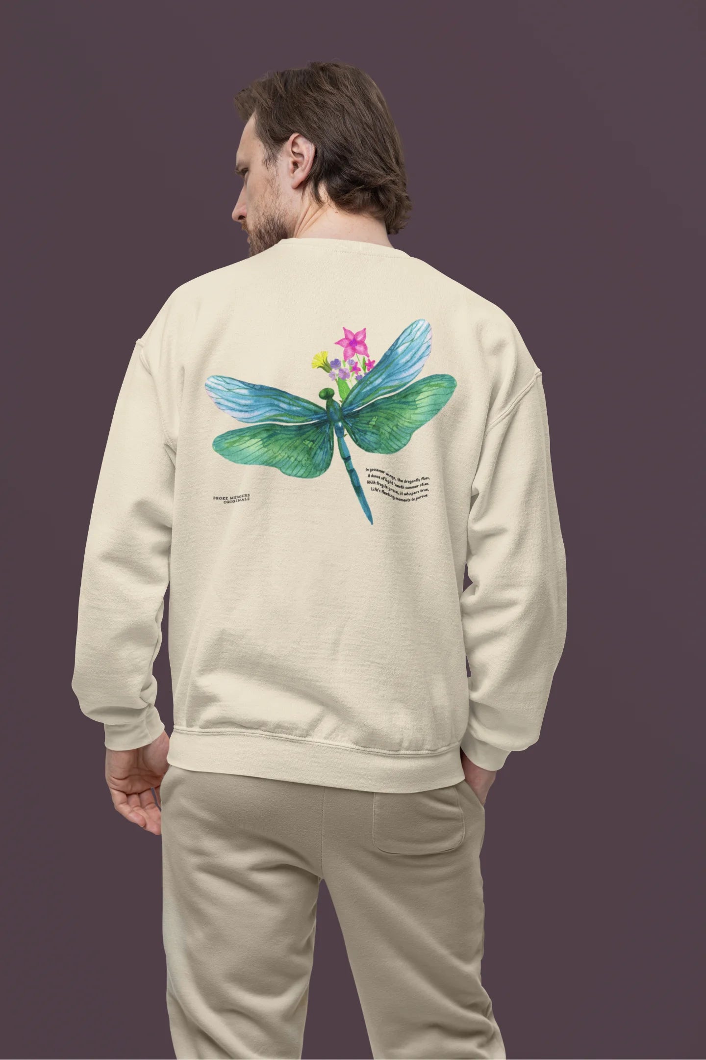 Back view of a male model wearing a cream sweatshirt featuring a nature-inspired design with a blue dragonfly perched on flowers amidst green grass. Ideal for those who love springtime aesthetics and dragonfly imagery.