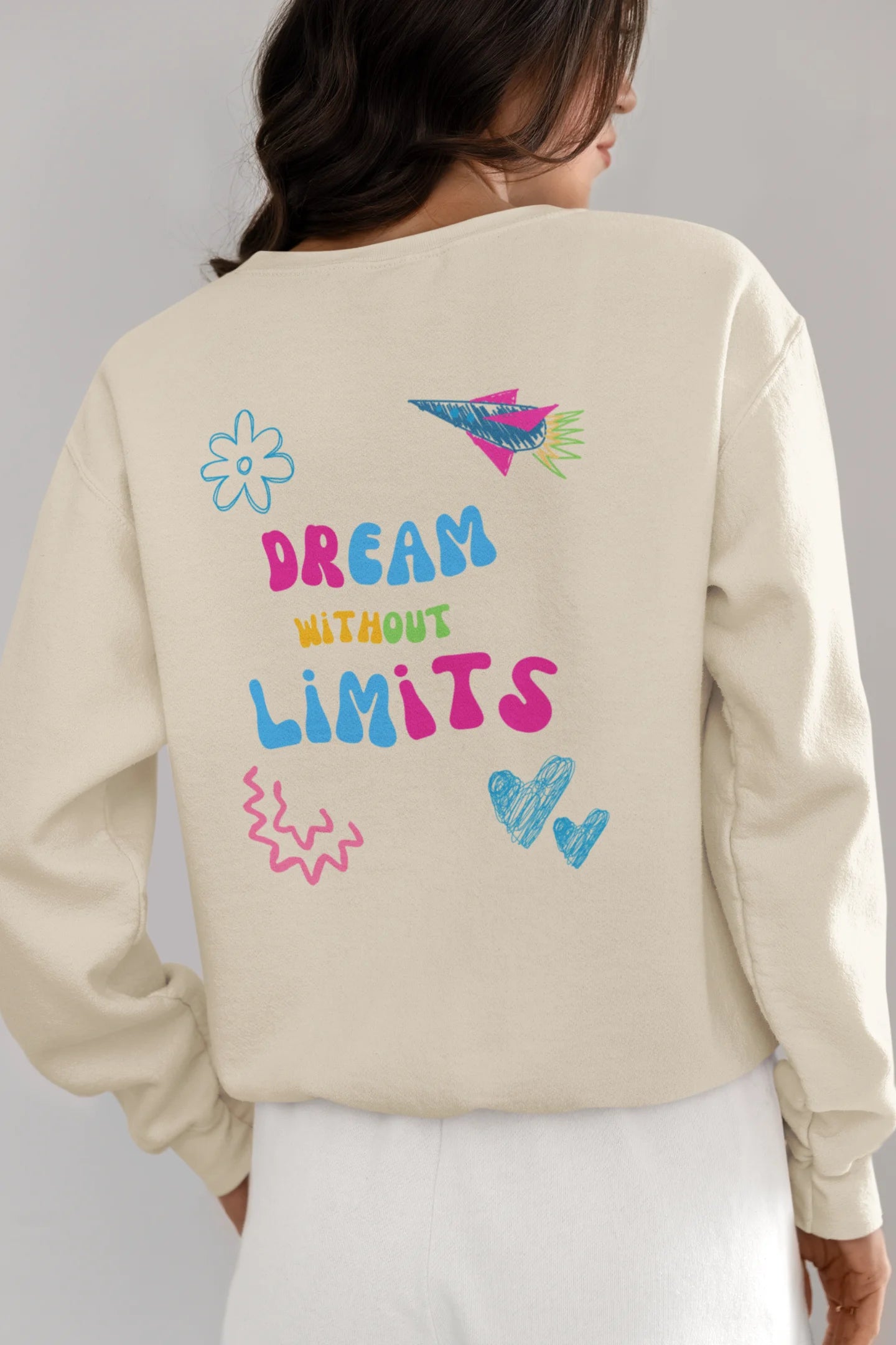 Back view of a female model wearing an cream sweatshirt featuring colorful "Dream Without Limits" text, offering an inspiring and uplifting aesthetic.