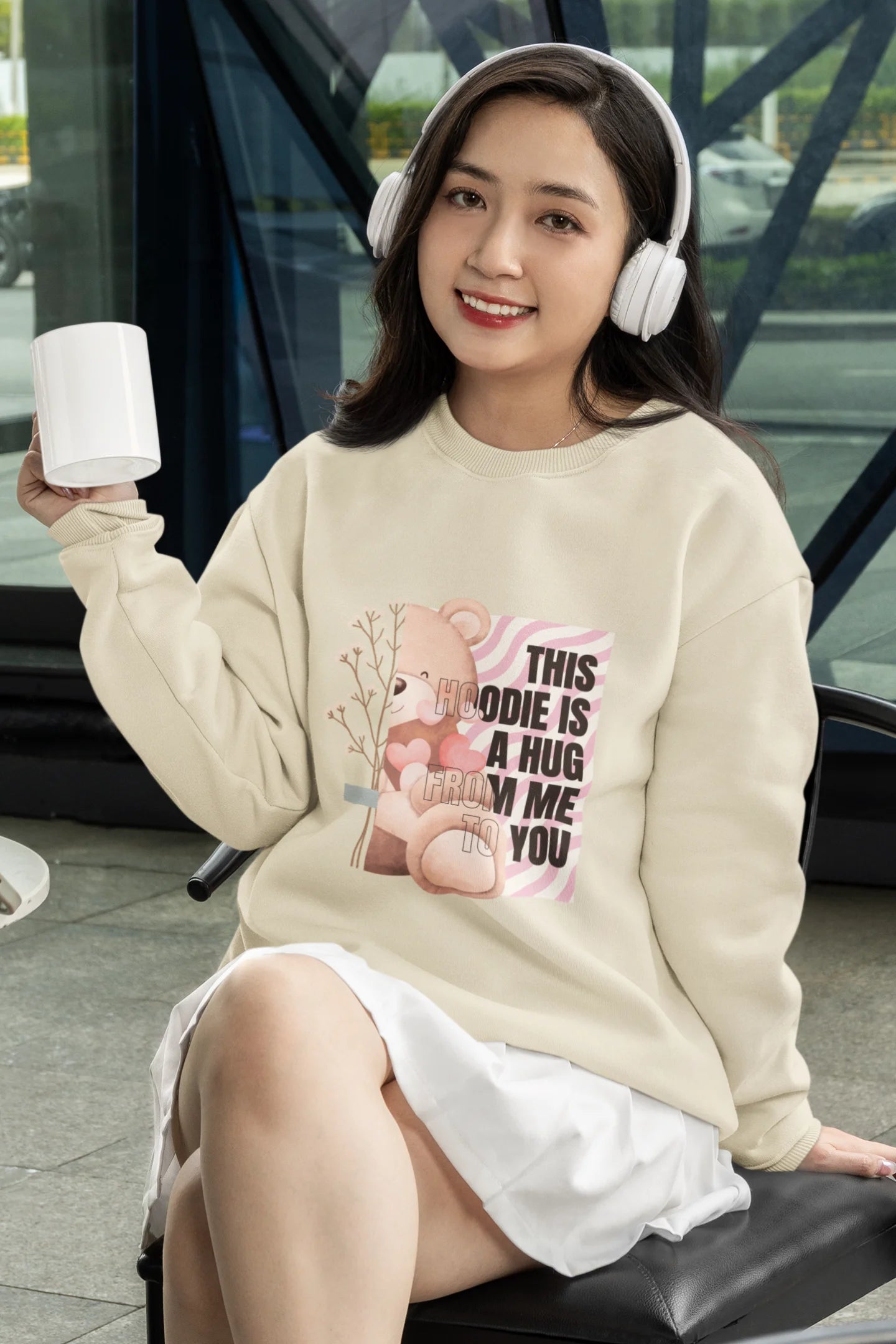 3rd front view of a female model wearing a cream sweatshirt with a heartwarming design of a teddy bear offering a hug, offering a comforting and relaxing aesthetic.
