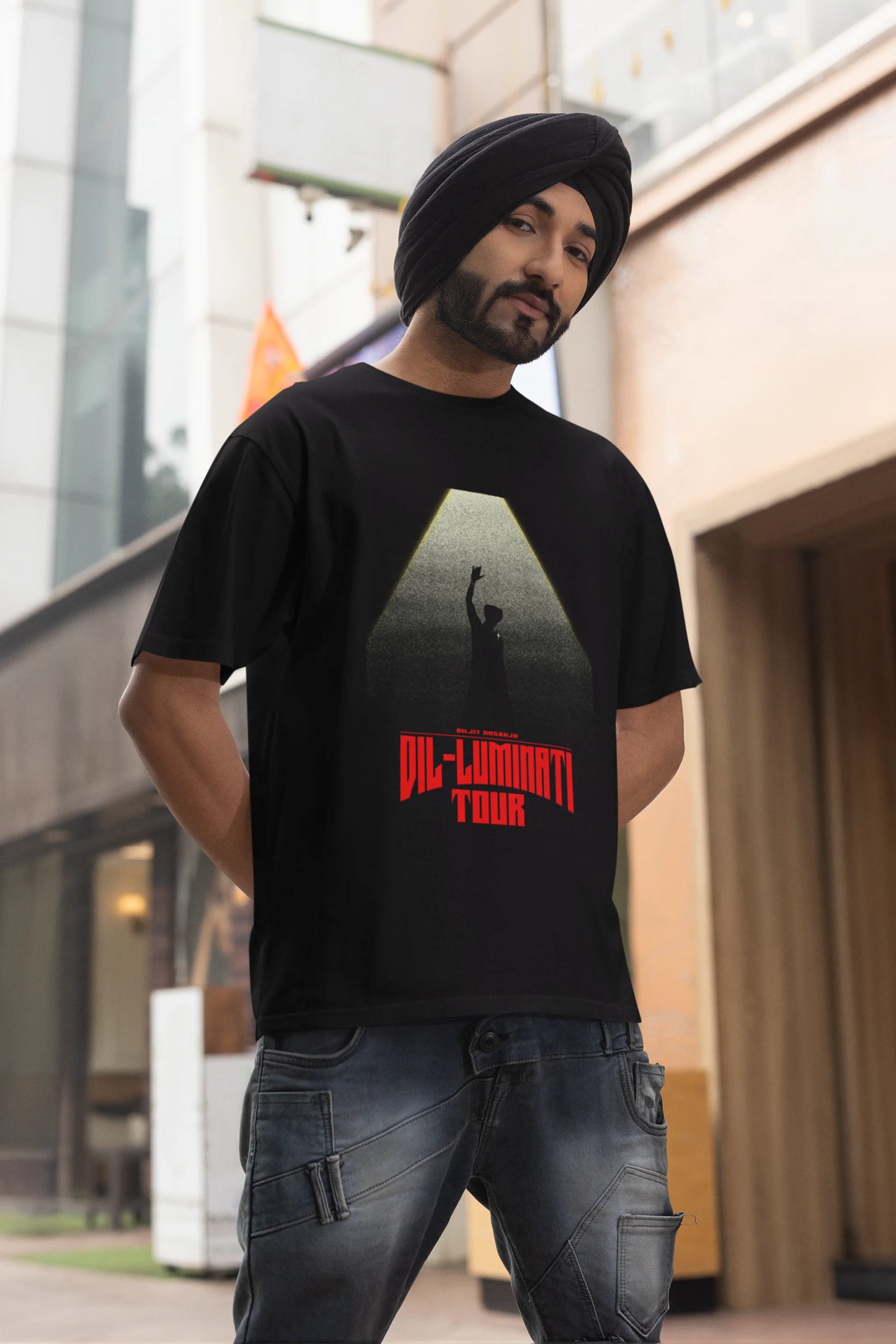 Fourth front view of a male model wearing a black oversized t-shirt with a portrait of Diljit Dosanjh on stage, bathed in the warm glow of a spotlight.