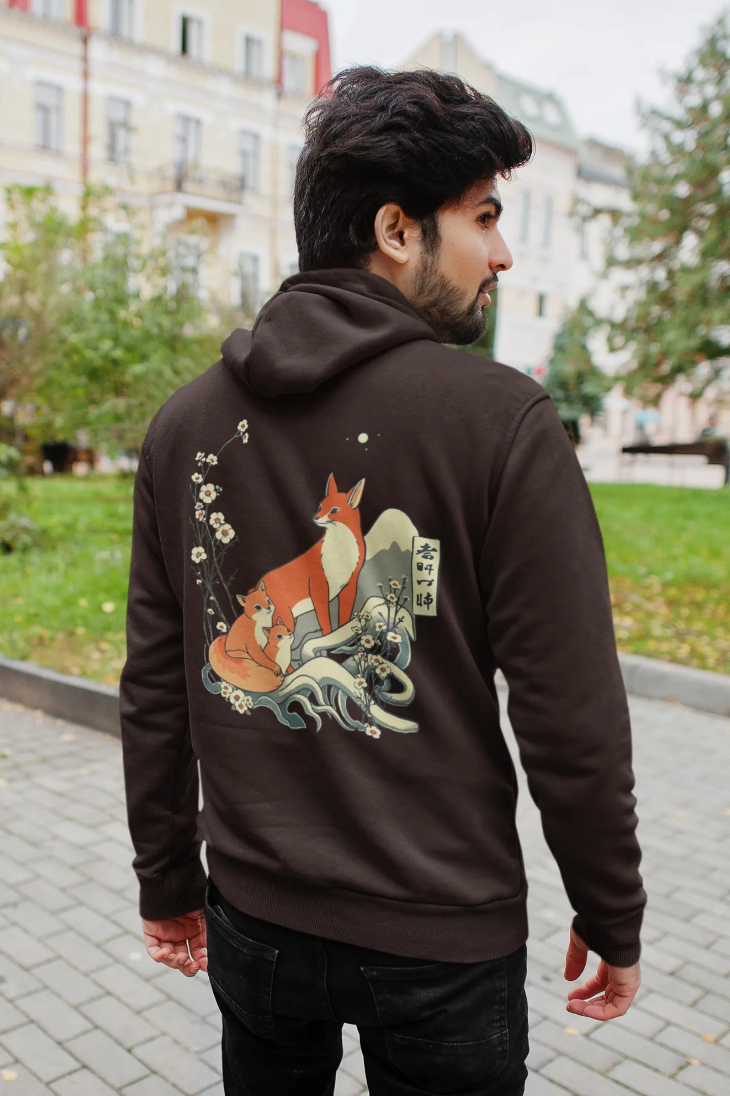 Back view male model wearing a chocolate brown hoodie with a design of a fox and its cub in a traditional Japanese landscape.