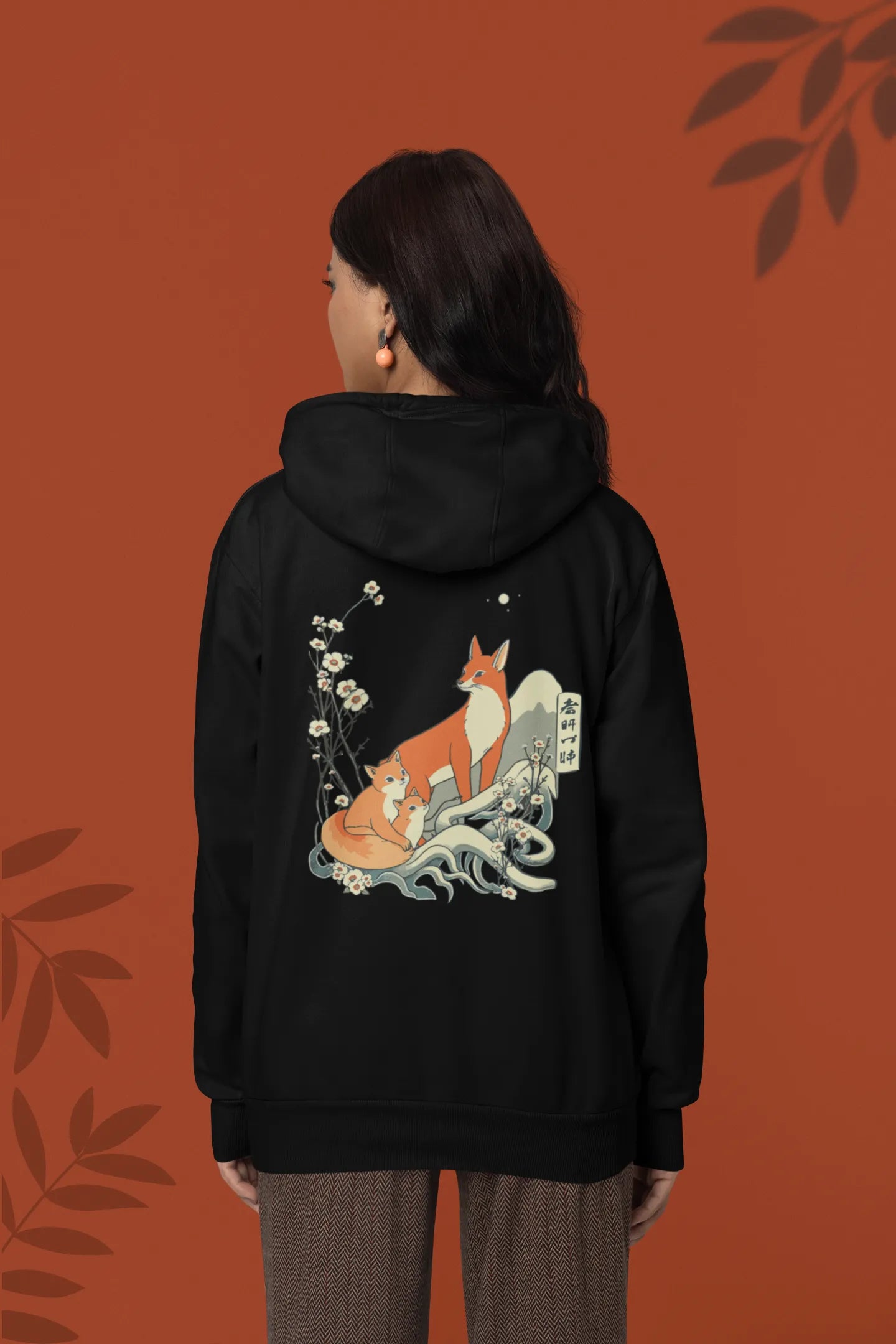 Back view of a female model wearing a black hoodie with a design of a fox and its cub in a traditional Japanese landscape.