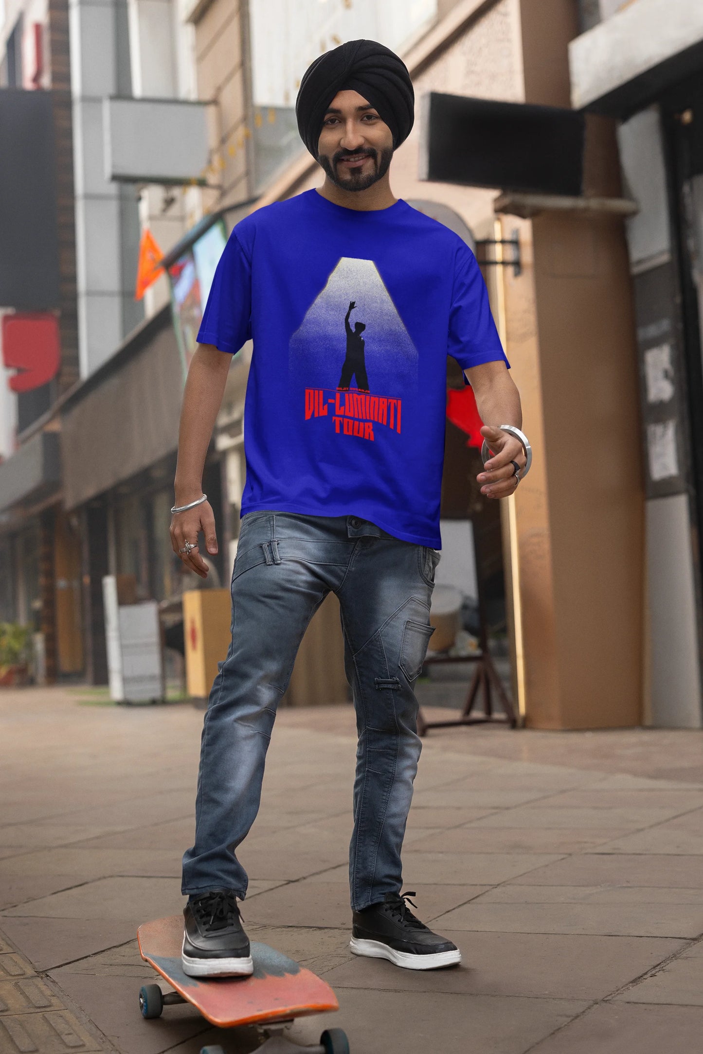 First front view of a male model wearing a royal blue oversized t-shirt with a portrait of Diljit Dosanjh on stage, bathed in the warm glow of a spotlight.