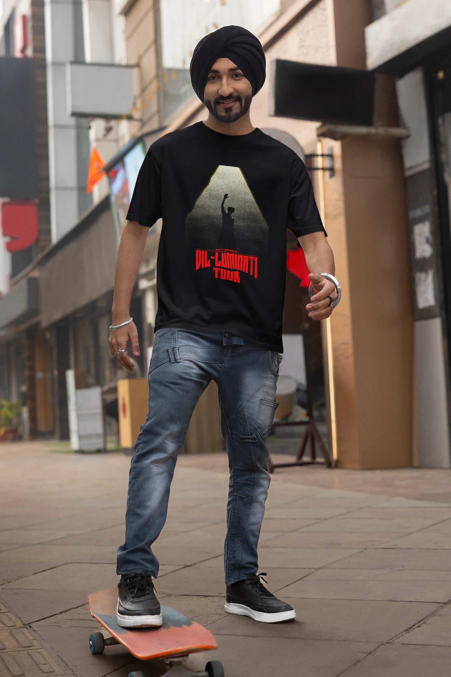 First front view of a male model wearing a black oversized t-shirt with a portrait of Diljit Dosanjh on stage, bathed in the warm glow of a spotlight.