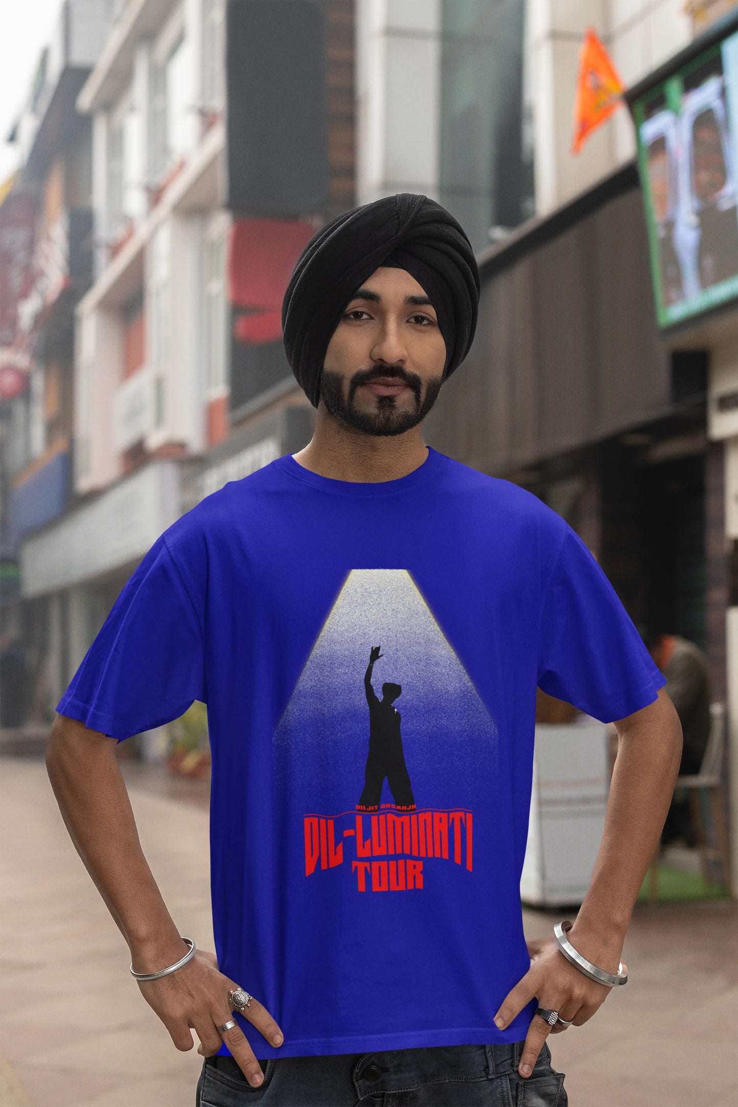 Third front view of a male model wearing a royal blue oversized t-shirt with a portrait of Diljit Dosanjh on stage, bathed in the warm glow of a spotlight.
