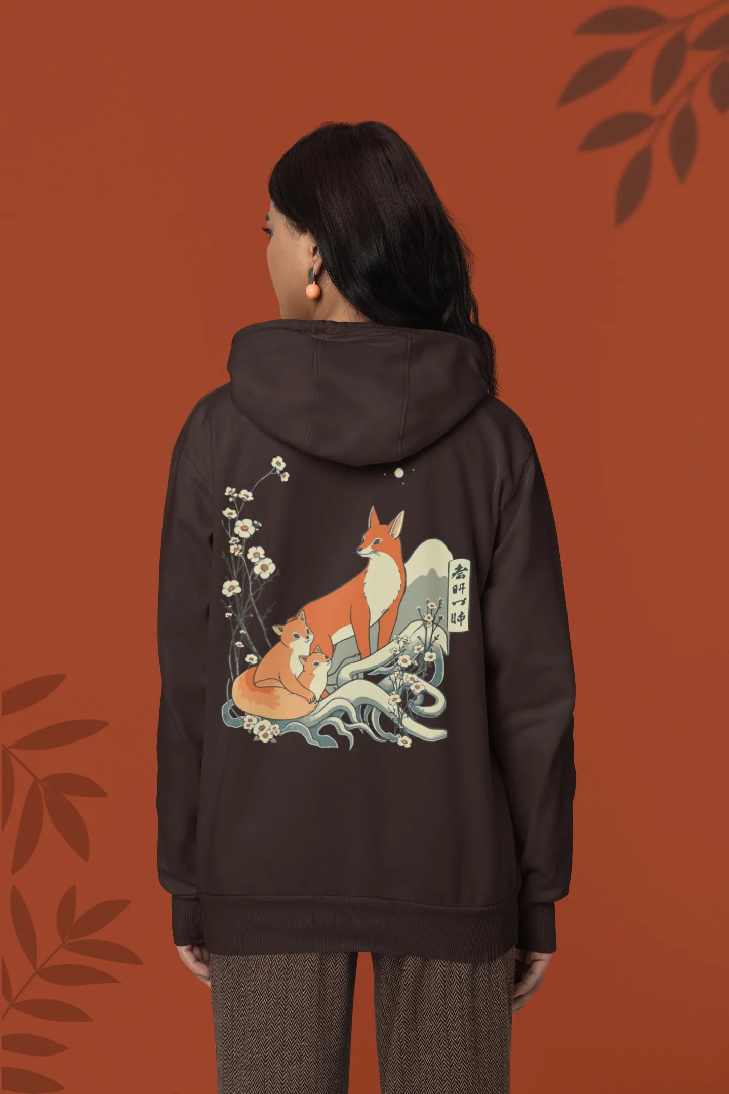Back view female model wearing a chocolate brown hoodie with a design of a fox and its cub in a traditional Japanese landscape.