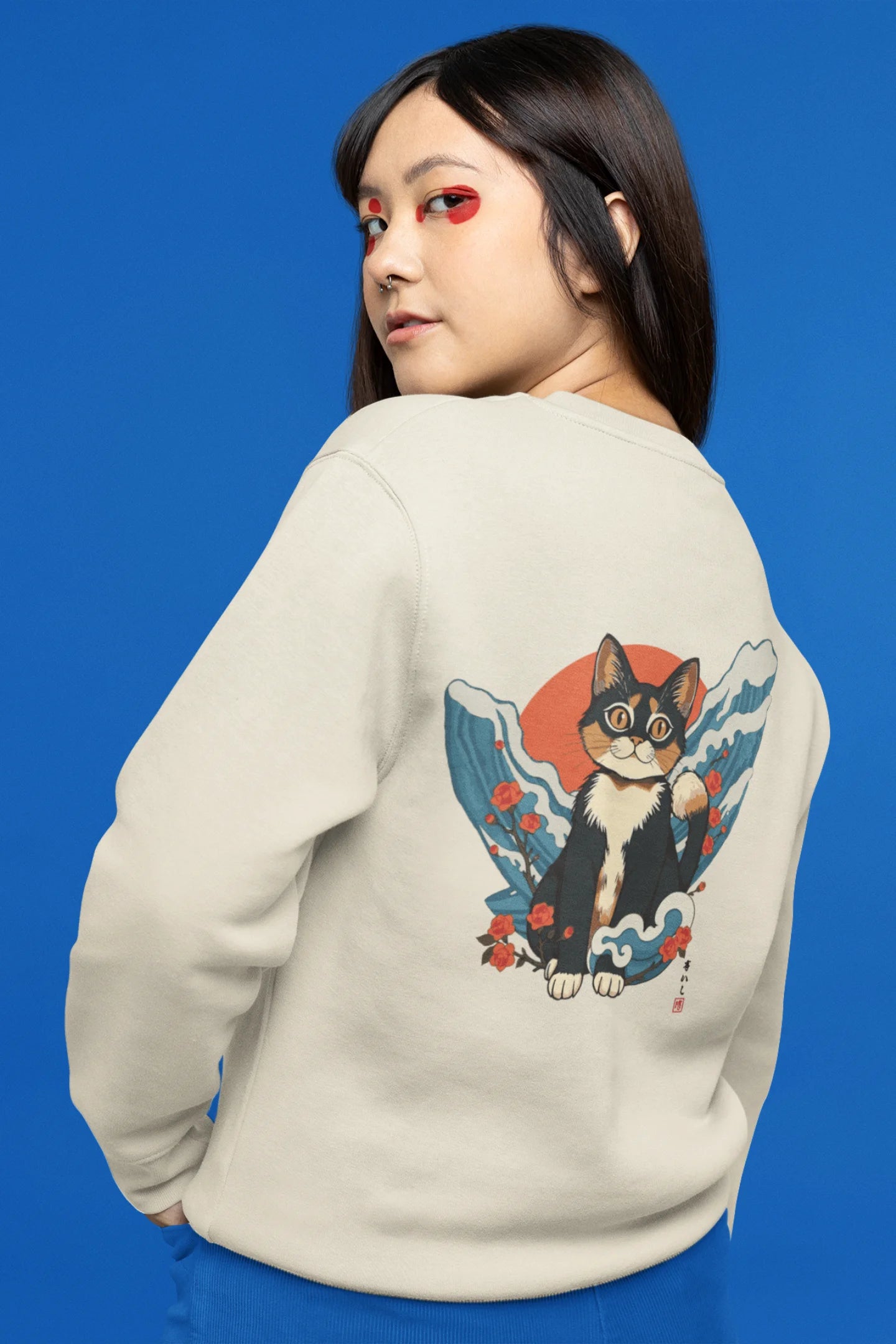 Back view of female model wearing a cream sweatshirt with a design of a cat dressed as a samurai, standing against a backdrop of waves and a rising sun.