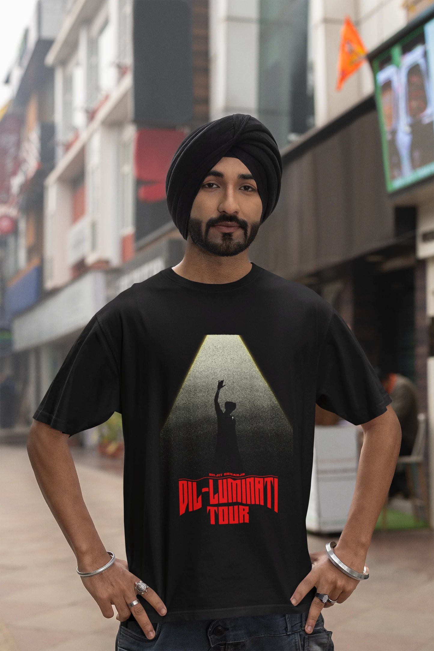 Third front view of a male model wearing a black oversized t-shirt with a portrait of Diljit Dosanjh on stage, bathed in the warm glow of a spotlight.