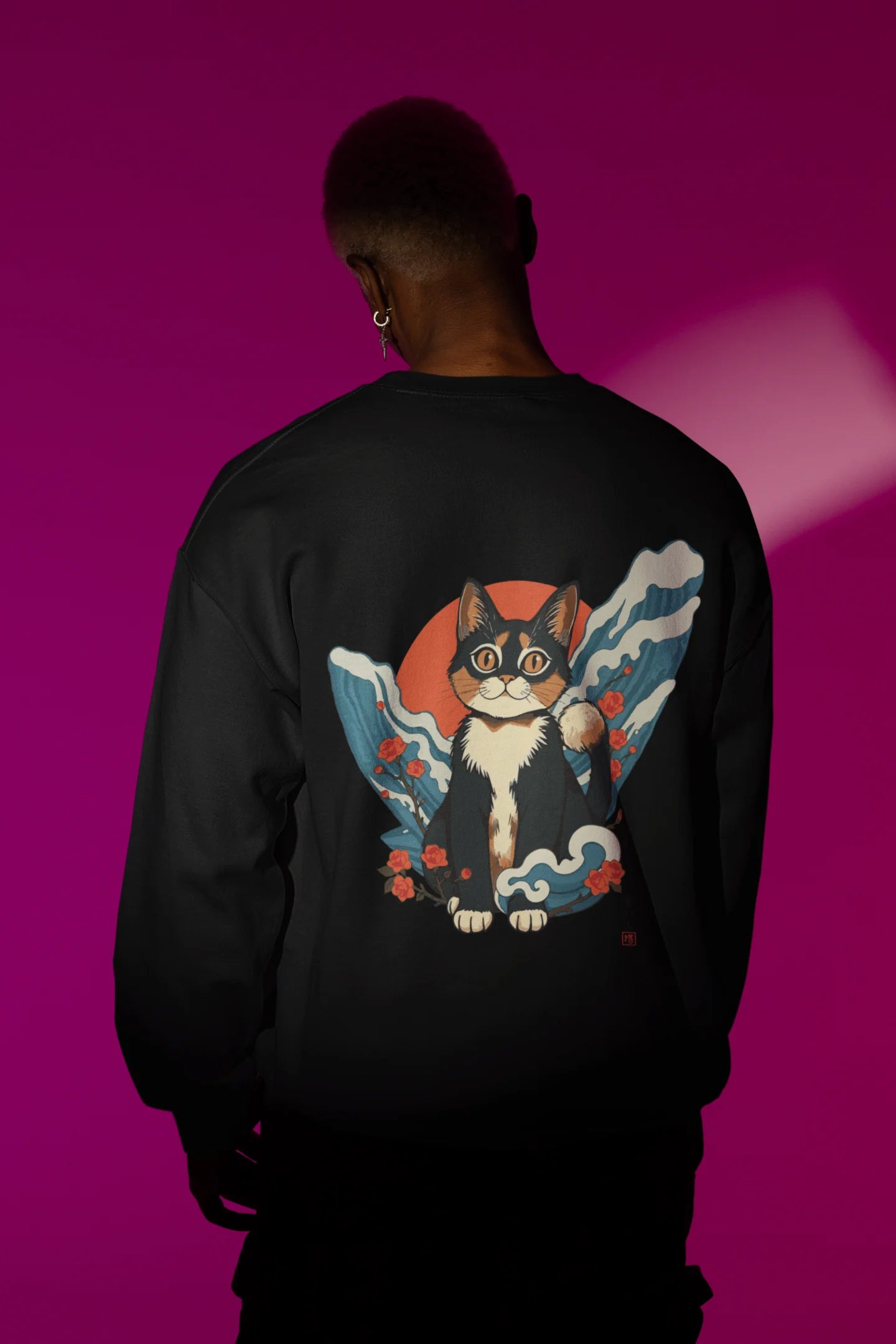 Back view of male model wearing a black sweatshirt with a design of a cat dressed as a samurai, standing against a backdrop of waves and a rising sun.