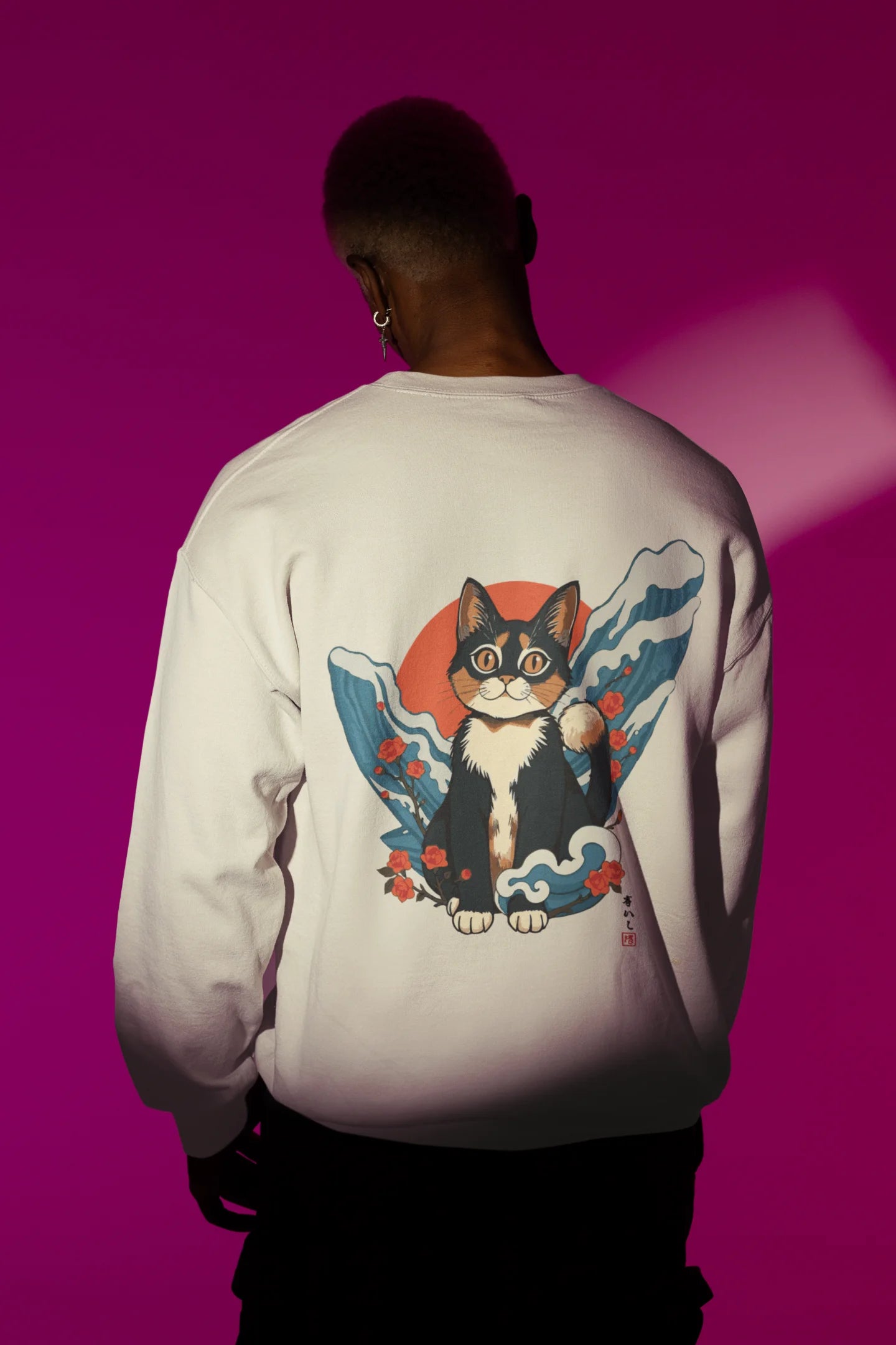 Back view of male model wearing a cream sweatshirt with a design of a cat dressed as a samurai, standing against a backdrop of waves and a rising sun.