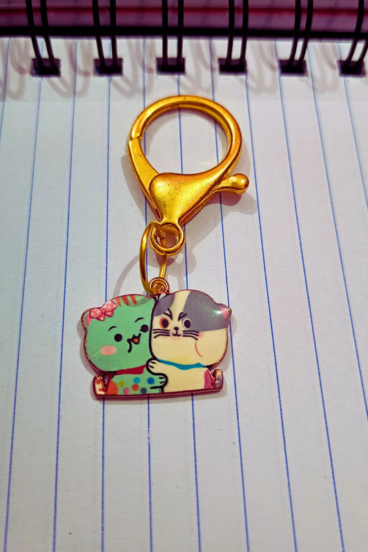 A view of a couples keychain featuring two separate charms depicting cats hugging each other. The cats' colors may vary, including combinations of blue and black and white.