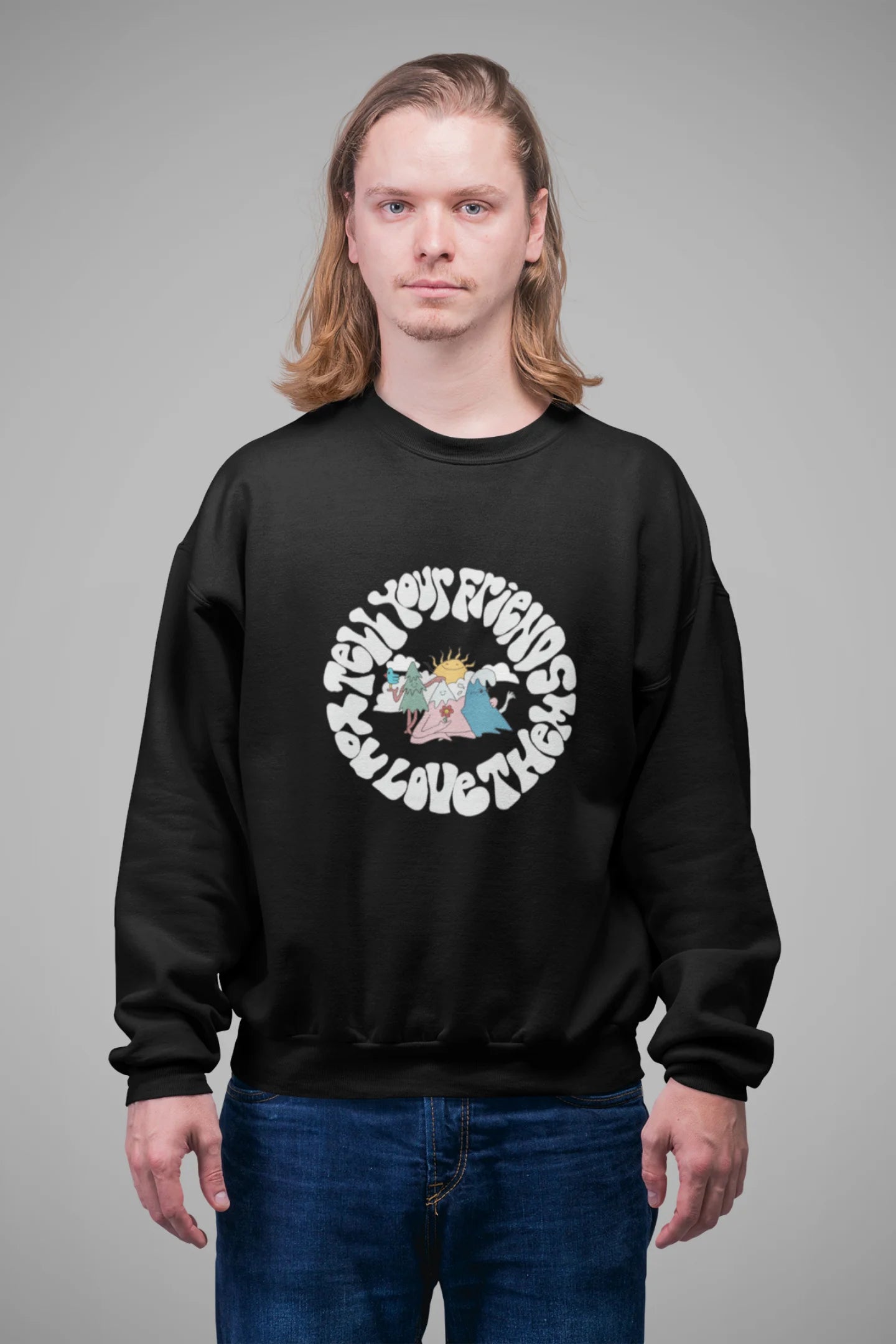 4th front view of a male model wearing a black sweatshirt with a design of smiling mountains and a sunrise, and the text "Tell Your Friends You Love Them."
