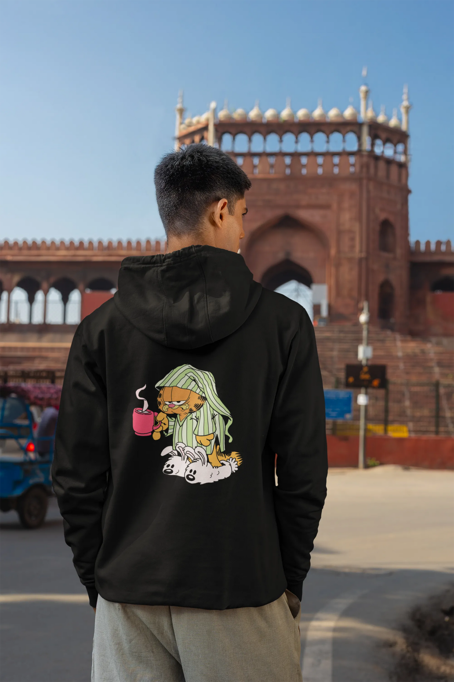 Back view of a male model wearing a black hoodie with a large Garfield design. The illustration shows Garfield standing out of bed, yawning and stretching, still wearing his nightcap and pajamas.