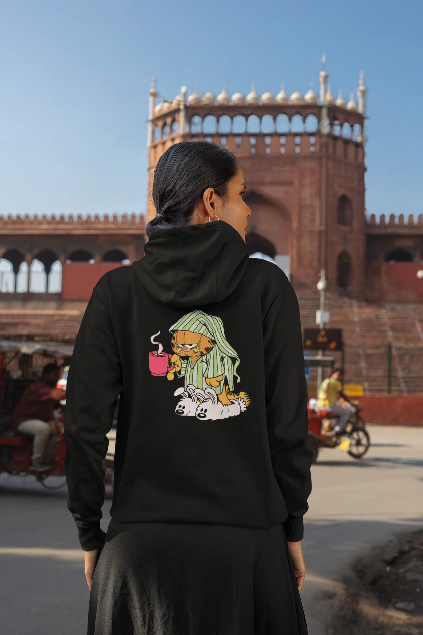Back view of a female model wearing a black hoodie with a large Garfield design. The illustration shows Garfield standing out of bed, yawning and stretching, still wearing his nightcap and pajamas.