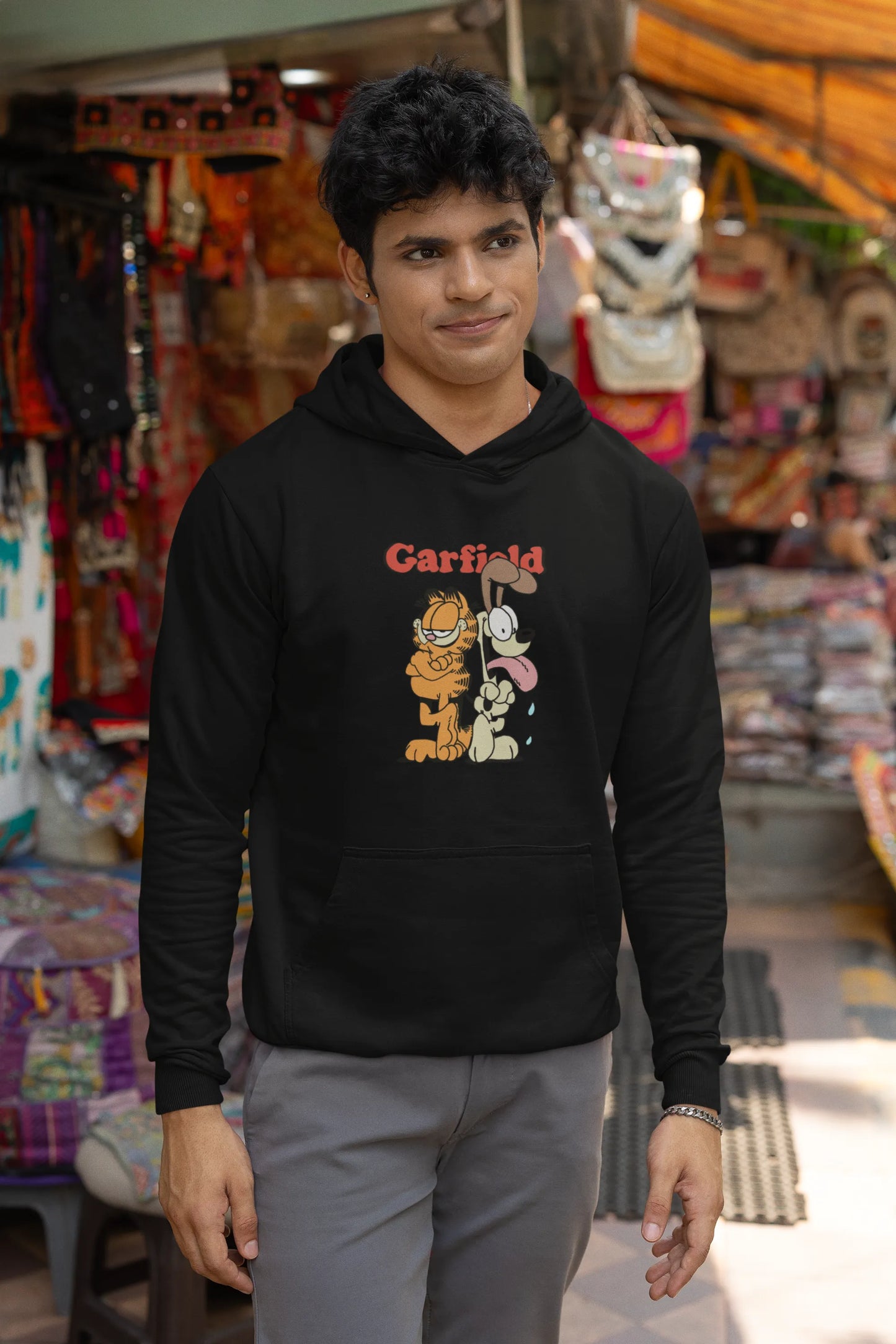 Second image of a male model wearing a black hoodie with a Garfield illustration showing the cat standing back to back with Odie, surrounded by a playful cartoon border.