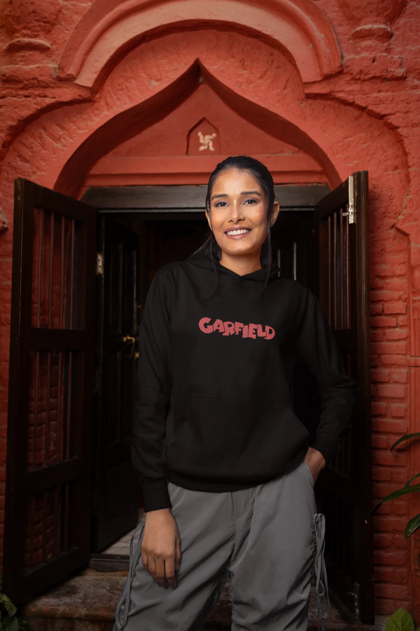 Second front view of a female model wearing a black hoodie with a large Garfield design. The illustration shows Garfield standing out of bed, yawning and stretching, still wearing his nightcap and pajamas.