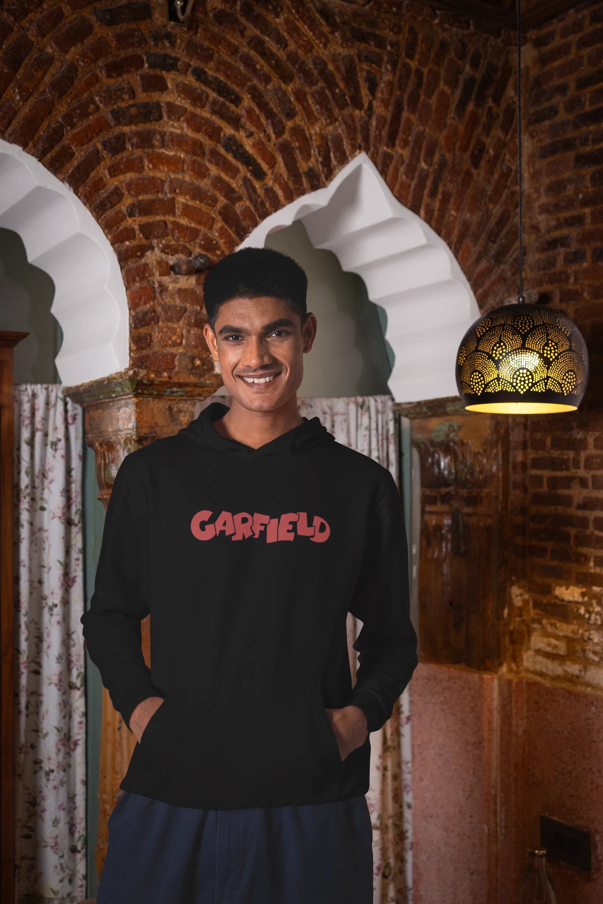 Third front view of a male model wearing a black hoodie with a large Garfield design. The illustration shows Garfield standing out of bed, yawning and stretching, still wearing his nightcap and pajamas.