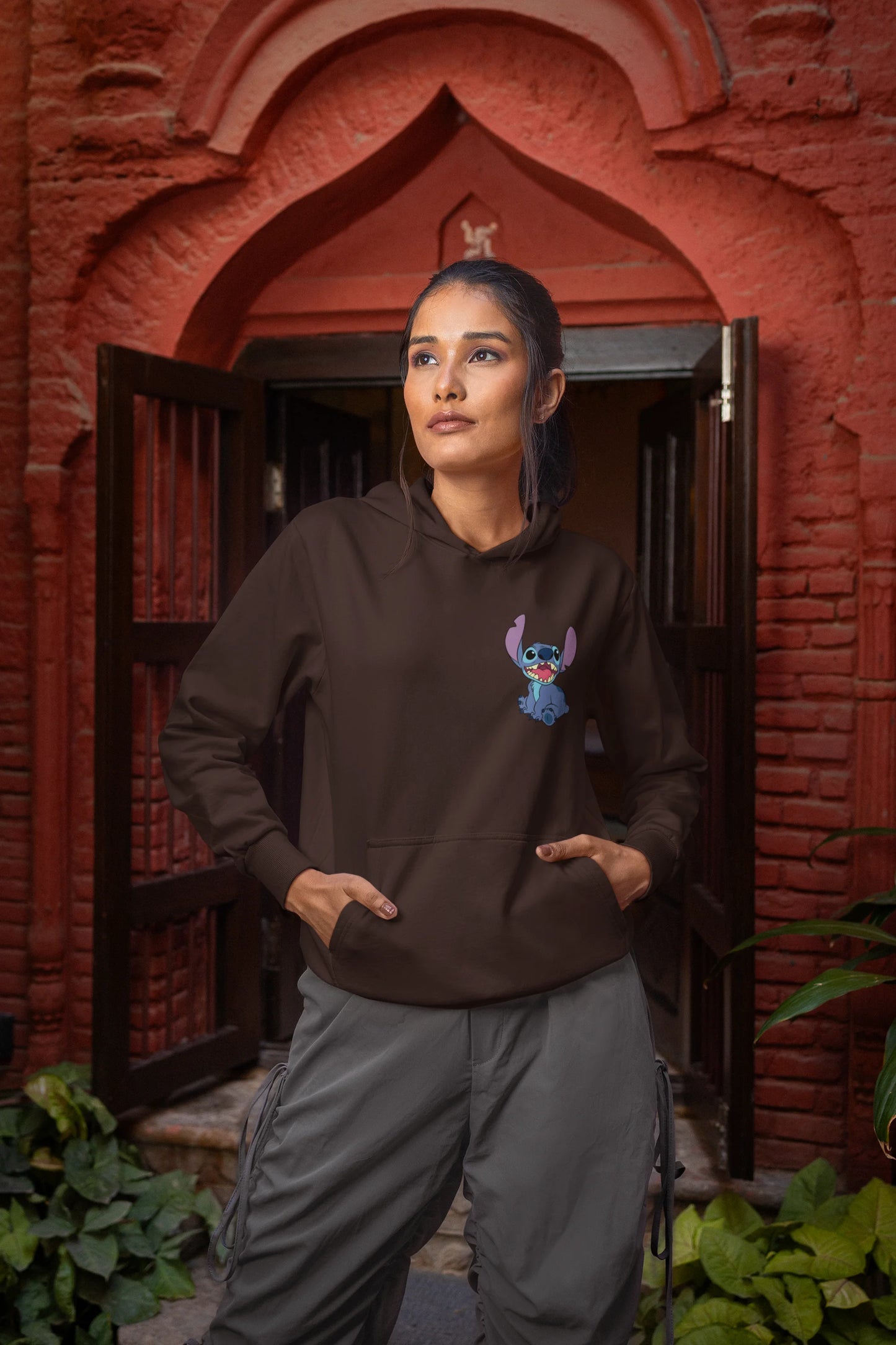 Front view of a female model wearing a chocolate brown hoodie with a collage of Stitch images. The illustration shows Stitch in different poses, making various funny faces and expressions.