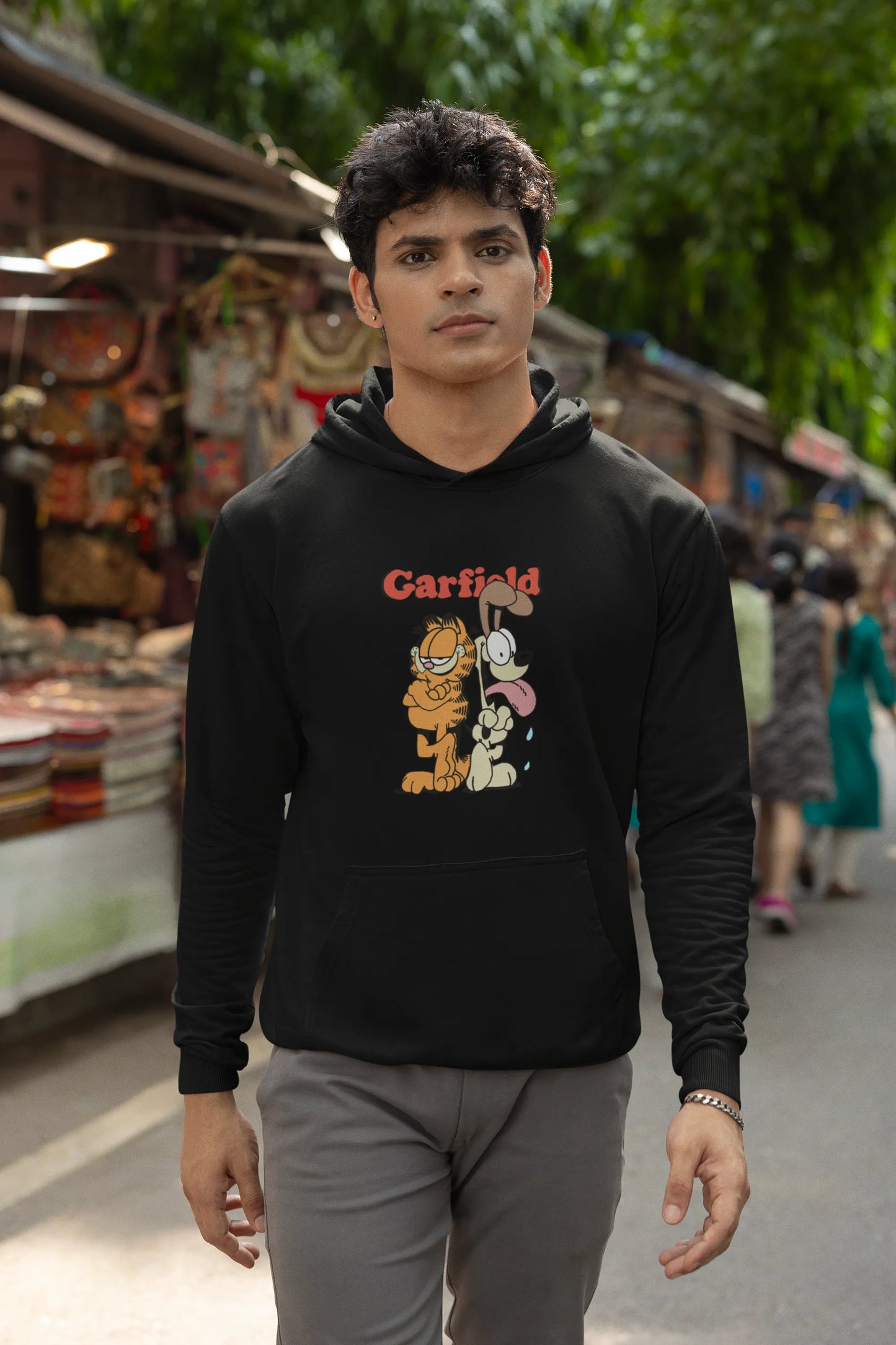 Fourth image of a male model wearing a black hoodie with a Garfield illustration showing the cat standing back to back with Odie, surrounded by a playful cartoon border.