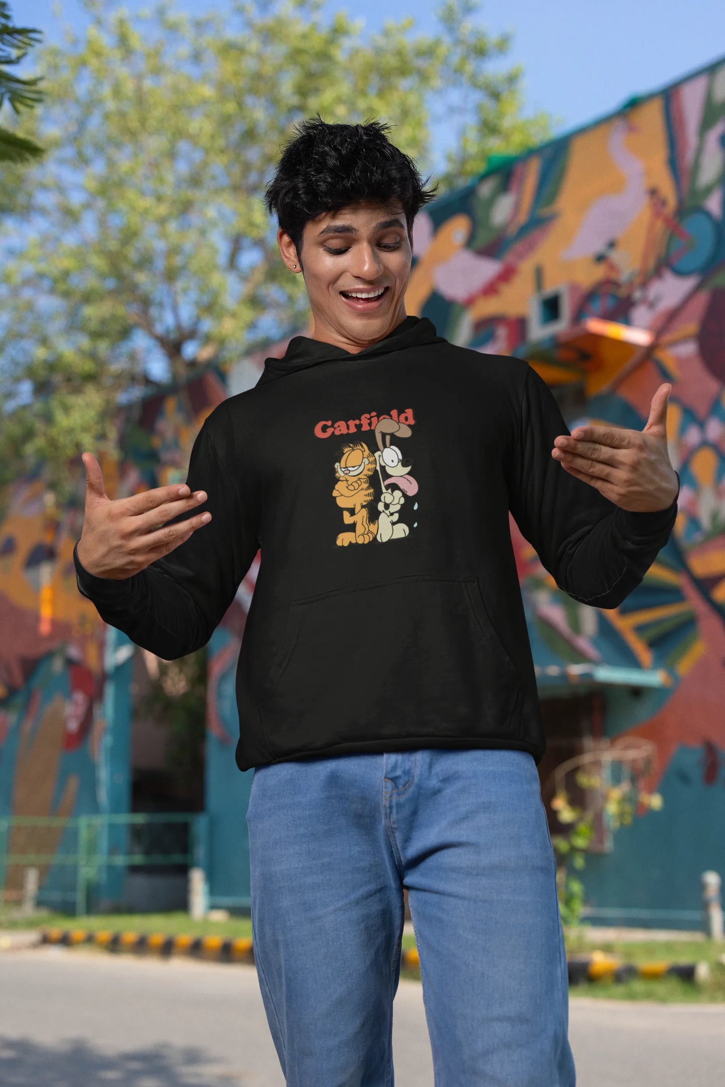 First image of a male model wearing a black hoodie with a Garfield illustration showing the cat standing back to back with Odie, surrounded by a playful cartoon border.