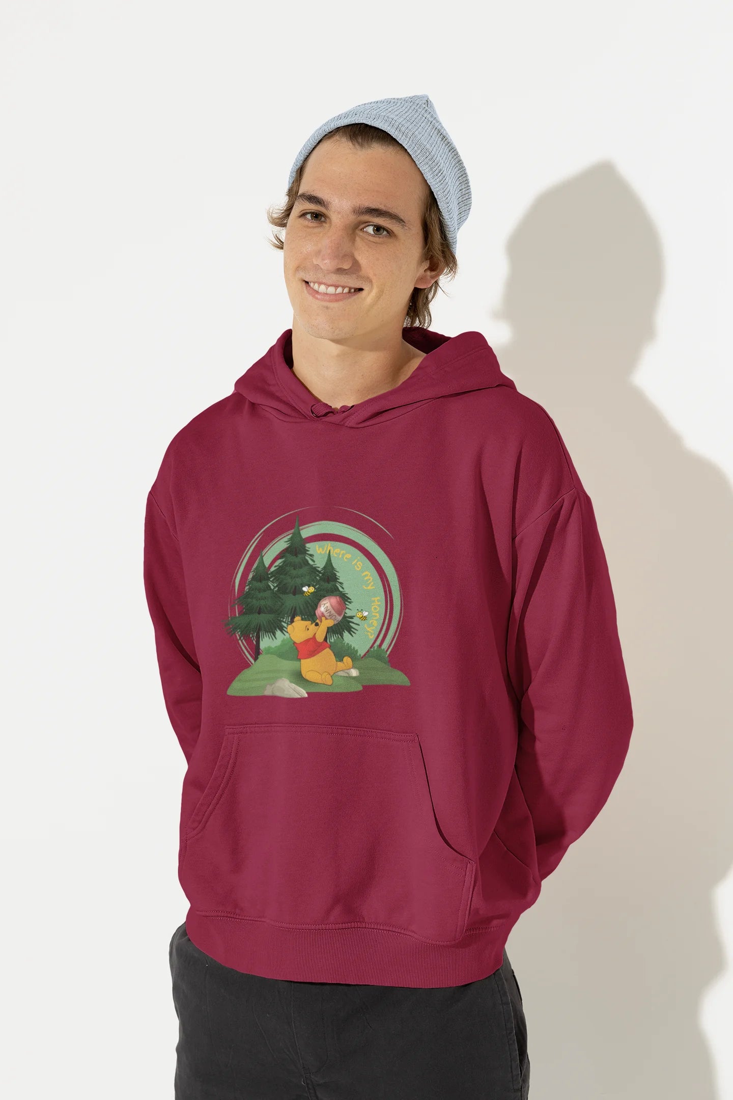 Front view of a male model wearing a maroon hoodie with a Winnie the Pooh design, showing the Disney bear eating honey from a jar in a forest.