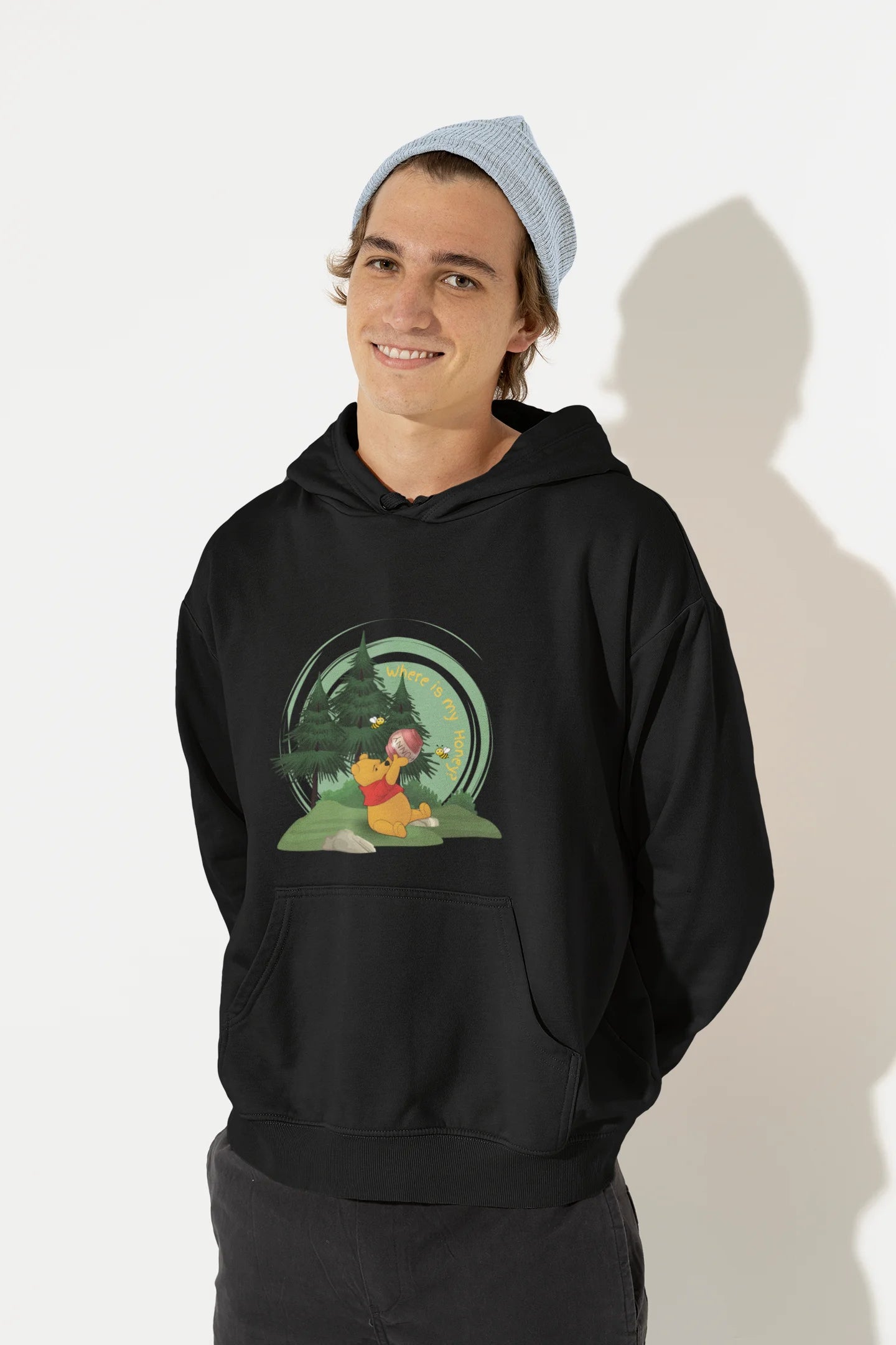 Front view of a male model wearing a black hoodie with a Winnie the Pooh design, showing the Disney bear eating honey from a jar in a forest.