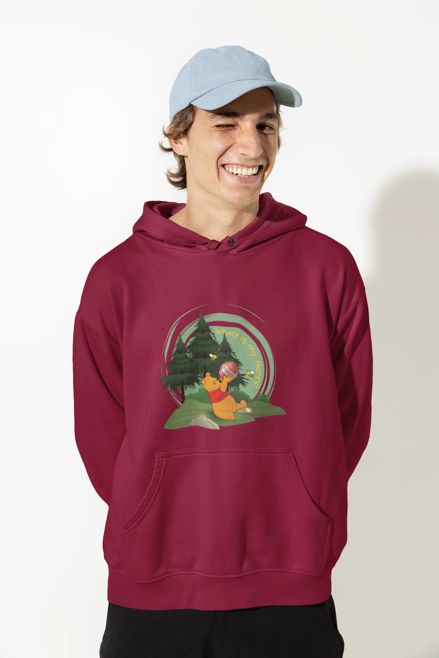 Close up front view of a male model wearing a maroon hoodie with a Winnie the Pooh design, showing the Disney bear eating honey from a jar in a forest.