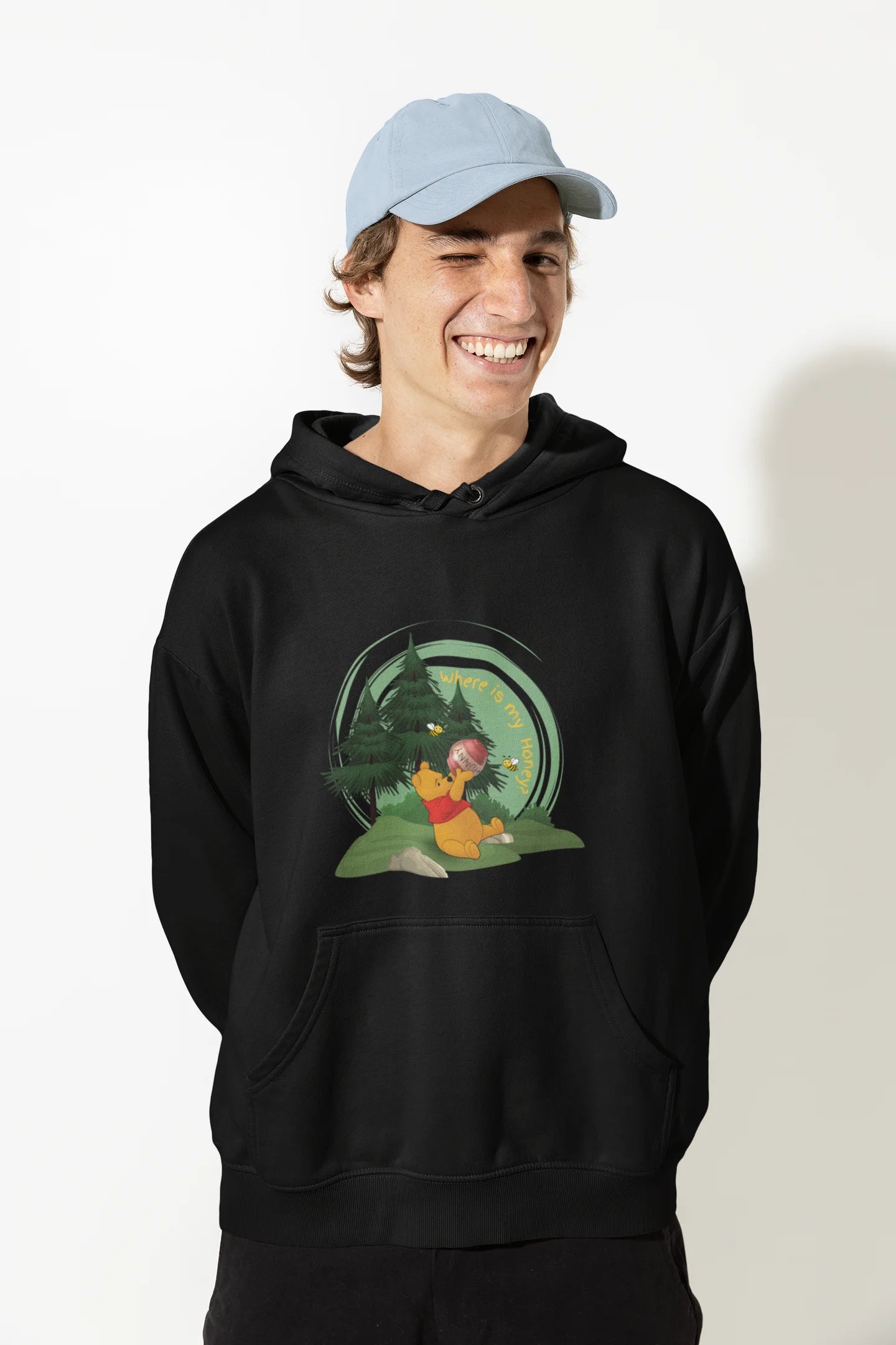 Close up front view of a male model wearing a black hoodie with a Winnie the Pooh design, showing the Disney bear eating honey from a jar in a forest.