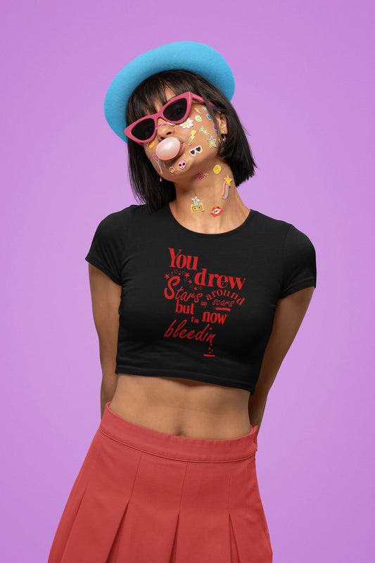 Second front view of a female model wearing a black crop top featuring the lyrics "You drew stars around my scars but now I'm bleeding," perfect for Swiftie fans.