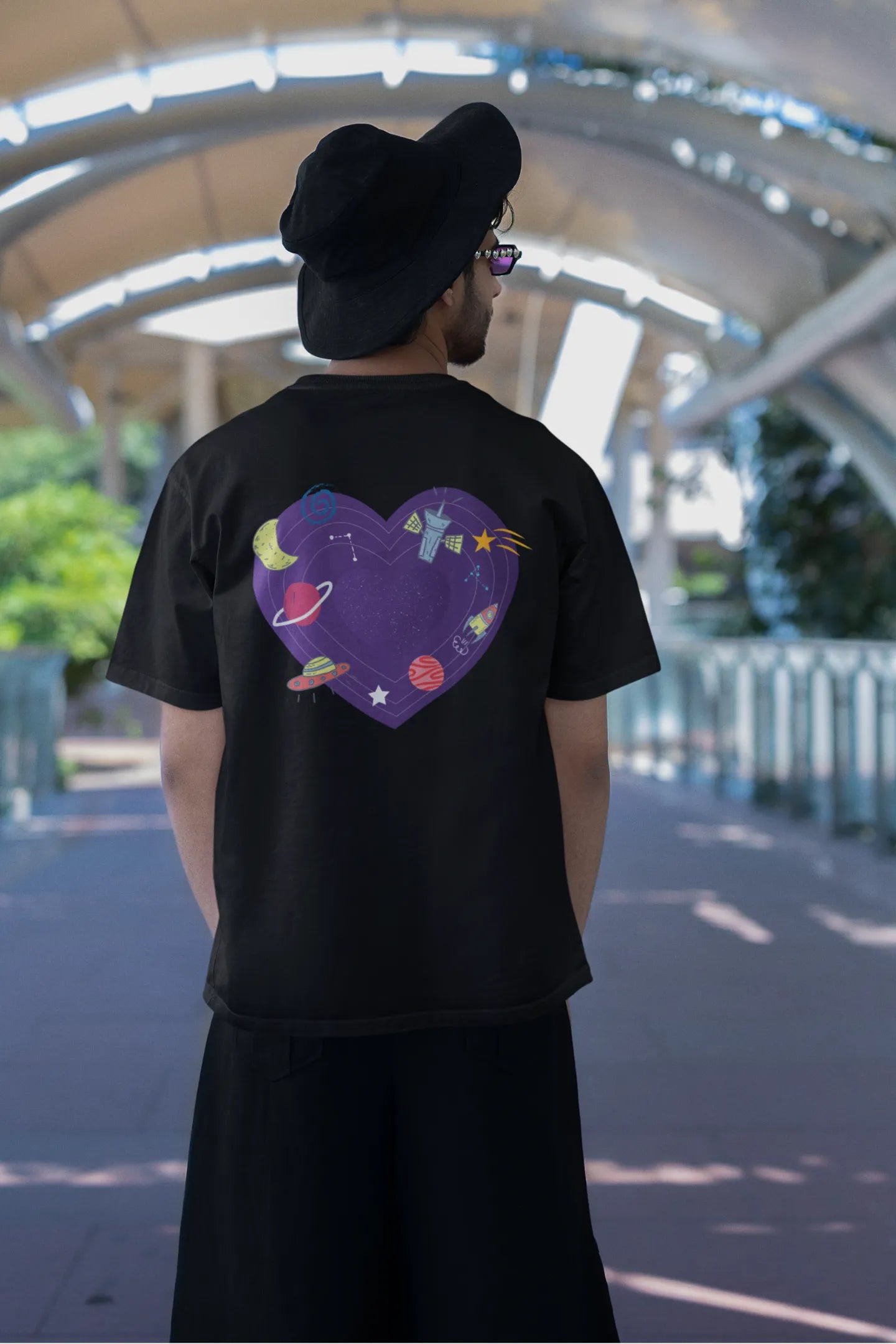 Back view of a male model wearing an black oversized t-shirt with the "Paradise" album artwork by Coldplay.
