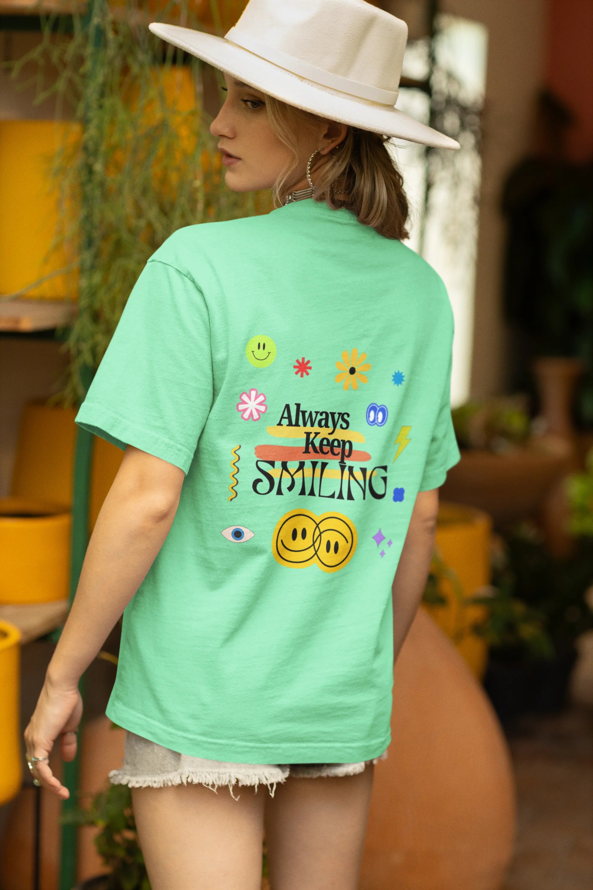 Back view of a female model wearing an oversized mint green t-shirt with a positive message "Keep Smiling" surrounded by colorful flowers and a smiley face emoji. Ideal for those who love cheerful clothing and spreading positivity.