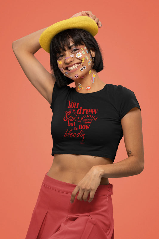 First front view of a female model wearing a black crop top featuring the lyrics "You drew stars around my scars but now I'm bleeding," perfect for Swiftie fans.