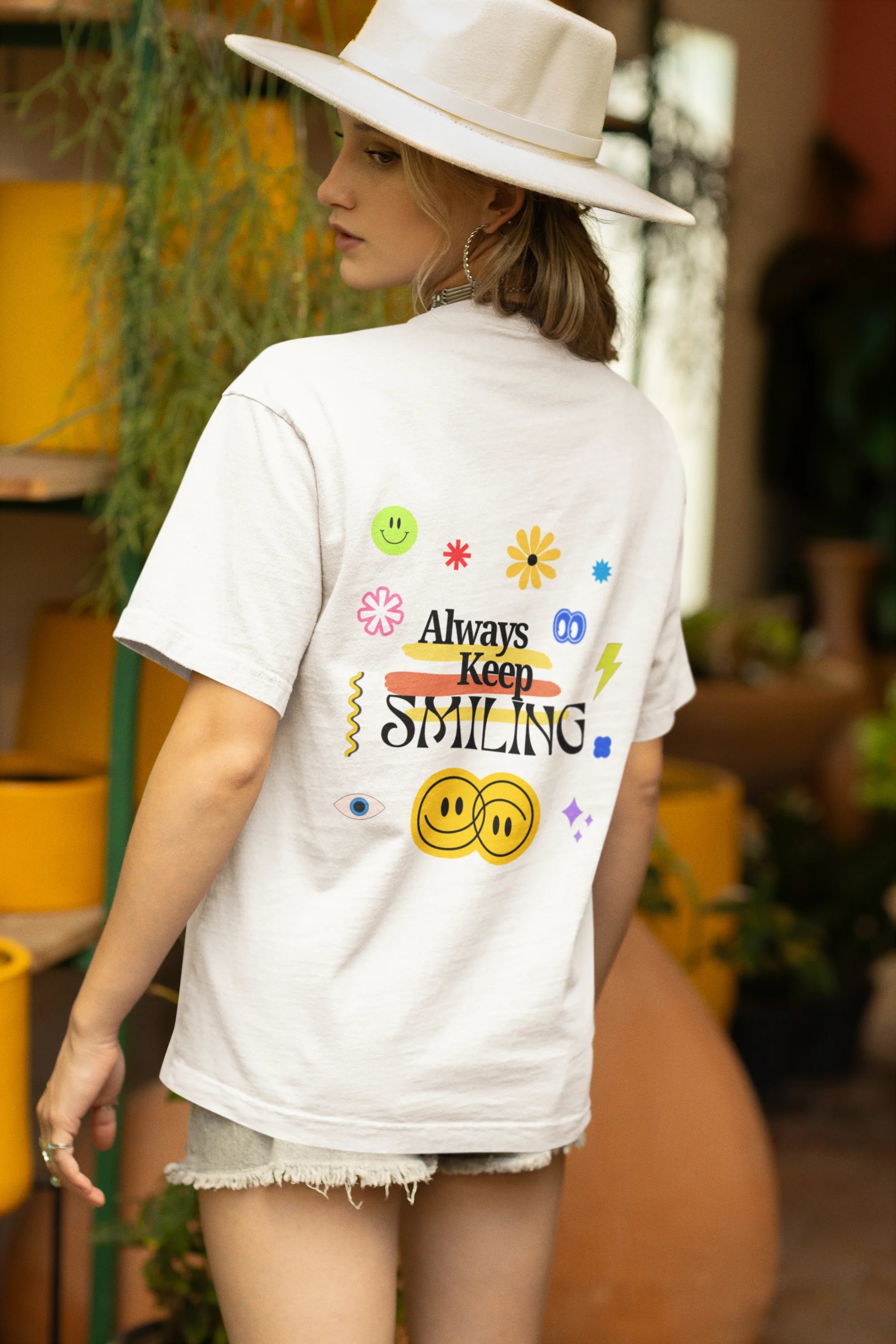 Back view of a female model wearing an oversized off-white t-shirt with a positive message "Keep Smiling" surrounded by colorful flowers and a smiley face emoji. Ideal for those who love cheerful clothing and spreading positivity.