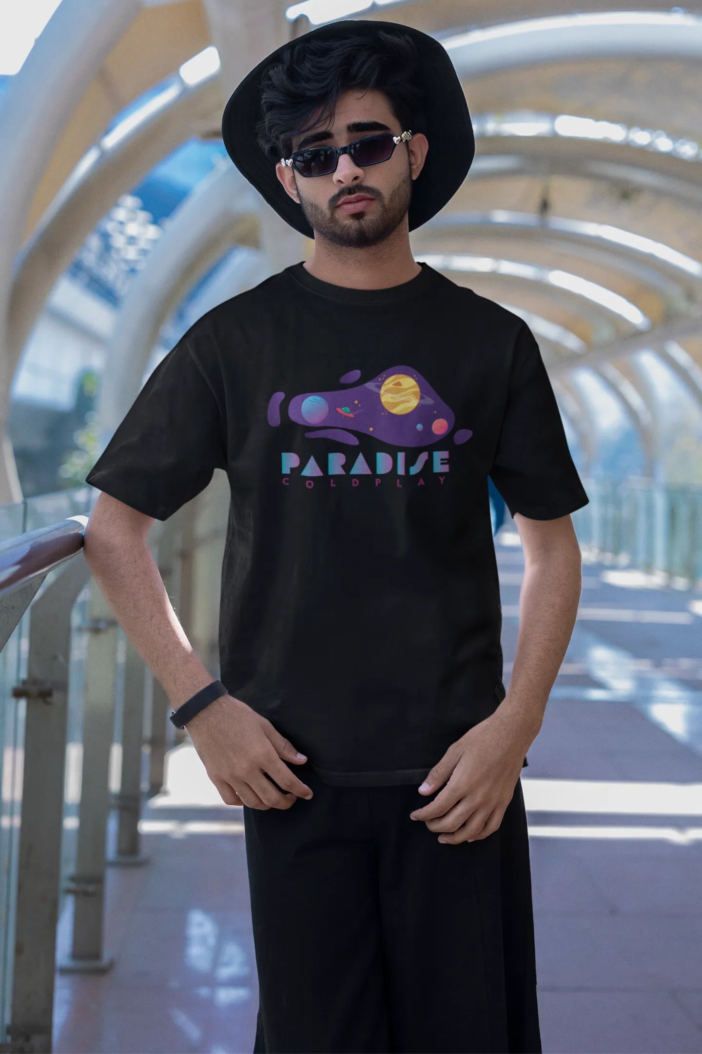 Second front view of a male model wearing an black oversized t-shirt with the "Paradise" album artwork by Coldplay.
