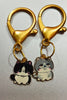 A couples keychain featuring two separate charms: one of a black cat and one of a white cat wearing glasses.