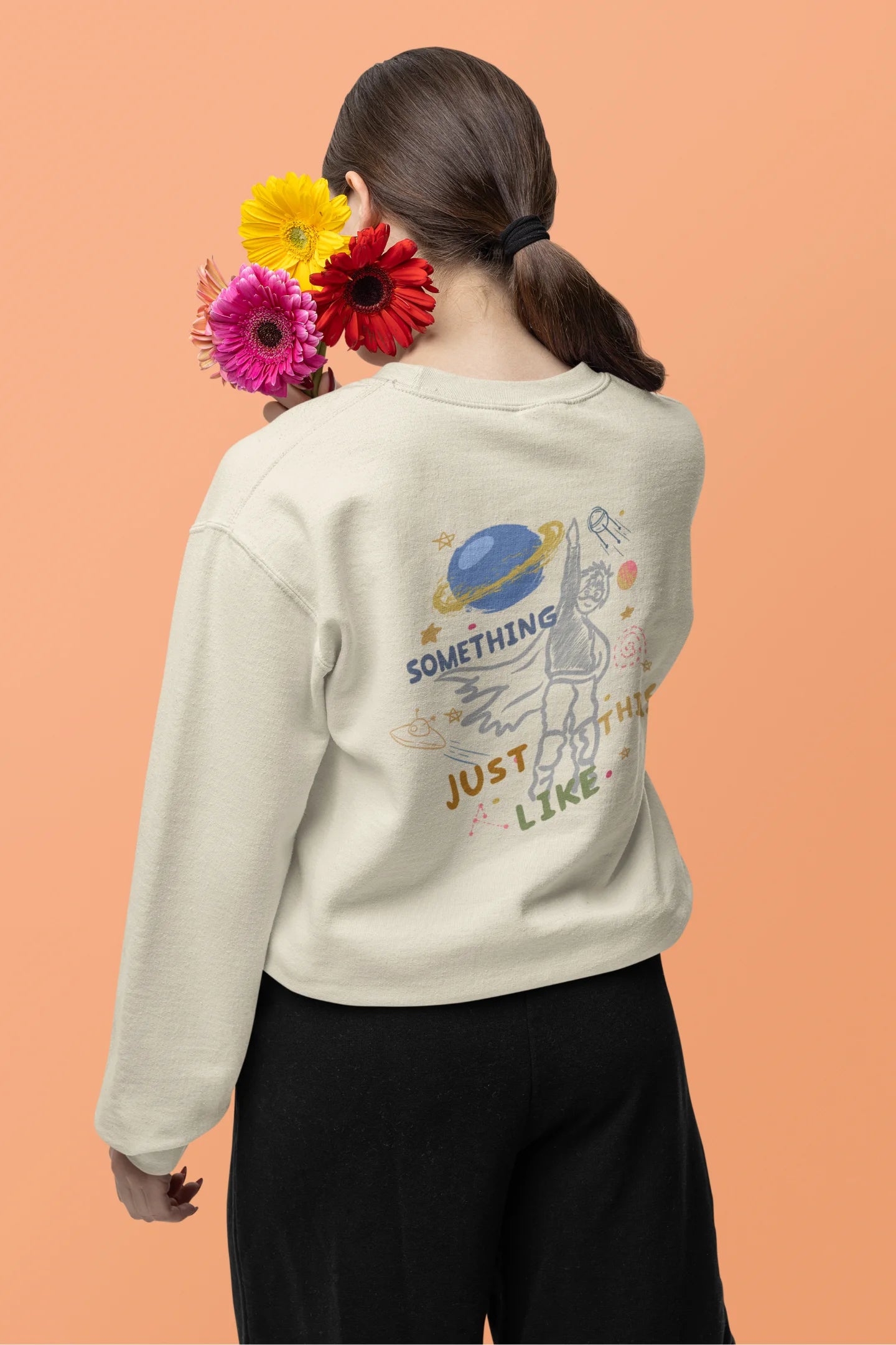 Back view of a female model wearing an cream sweatshirt with a design inspired by the "Something Just Like This" music video, featuring the word "Superheroes".