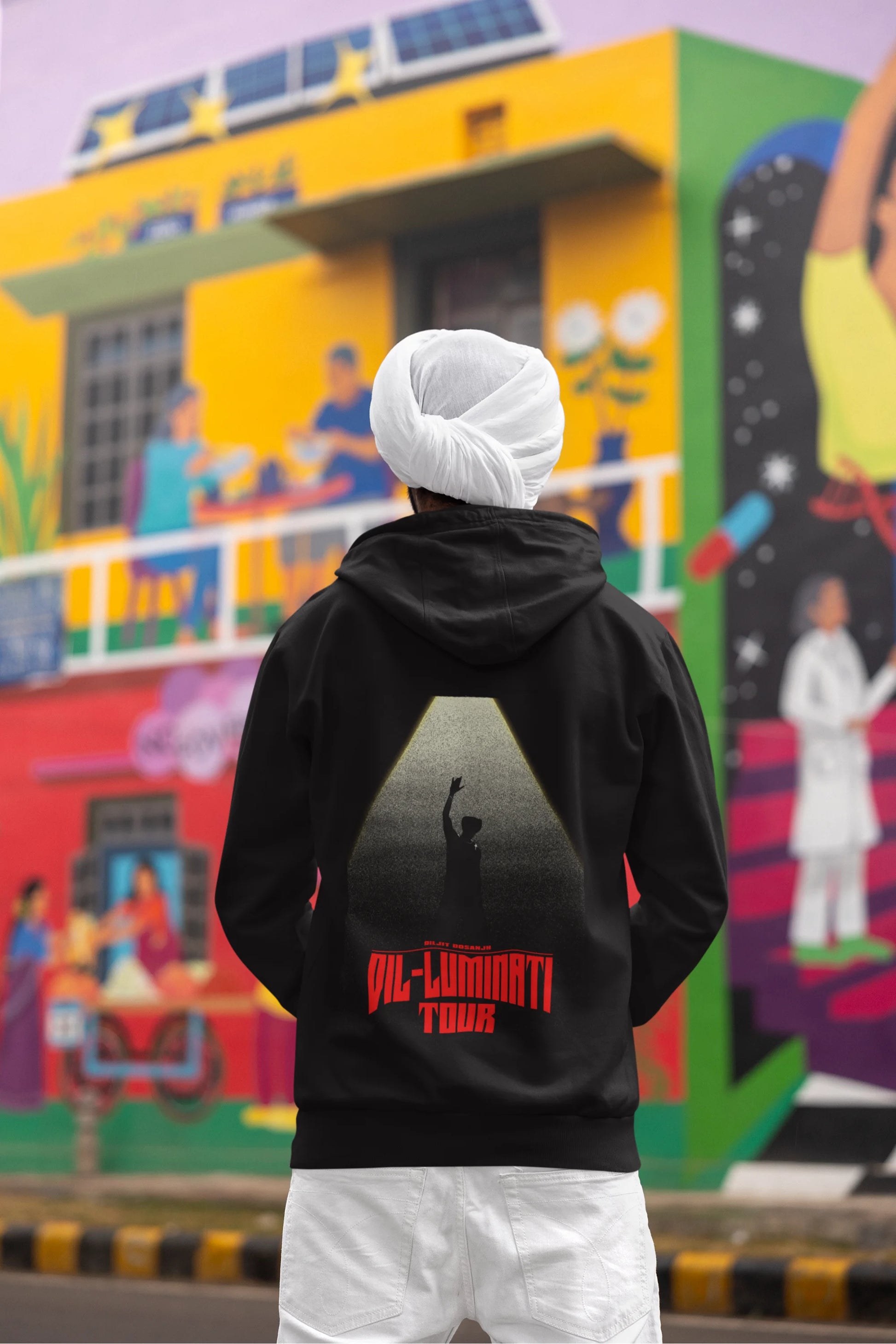 Back view of a male model wearing a black hoodie with a portrait of Diljit Dosanjh on stage, illuminated by a spotlight.