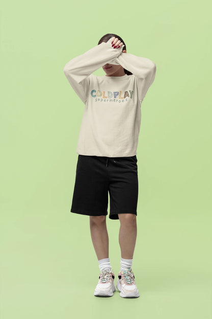 Fourth front view of a female model wearing an cream sweatshirt with a design inspired by the "Something Just Like This" music video, featuring the word "Superheroes".
