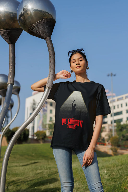 First front view of a woman model wearing a black oversized t-shirt with a portrait of Diljit Dosanjh on stage, bathed in the warm glow of a spotlight.