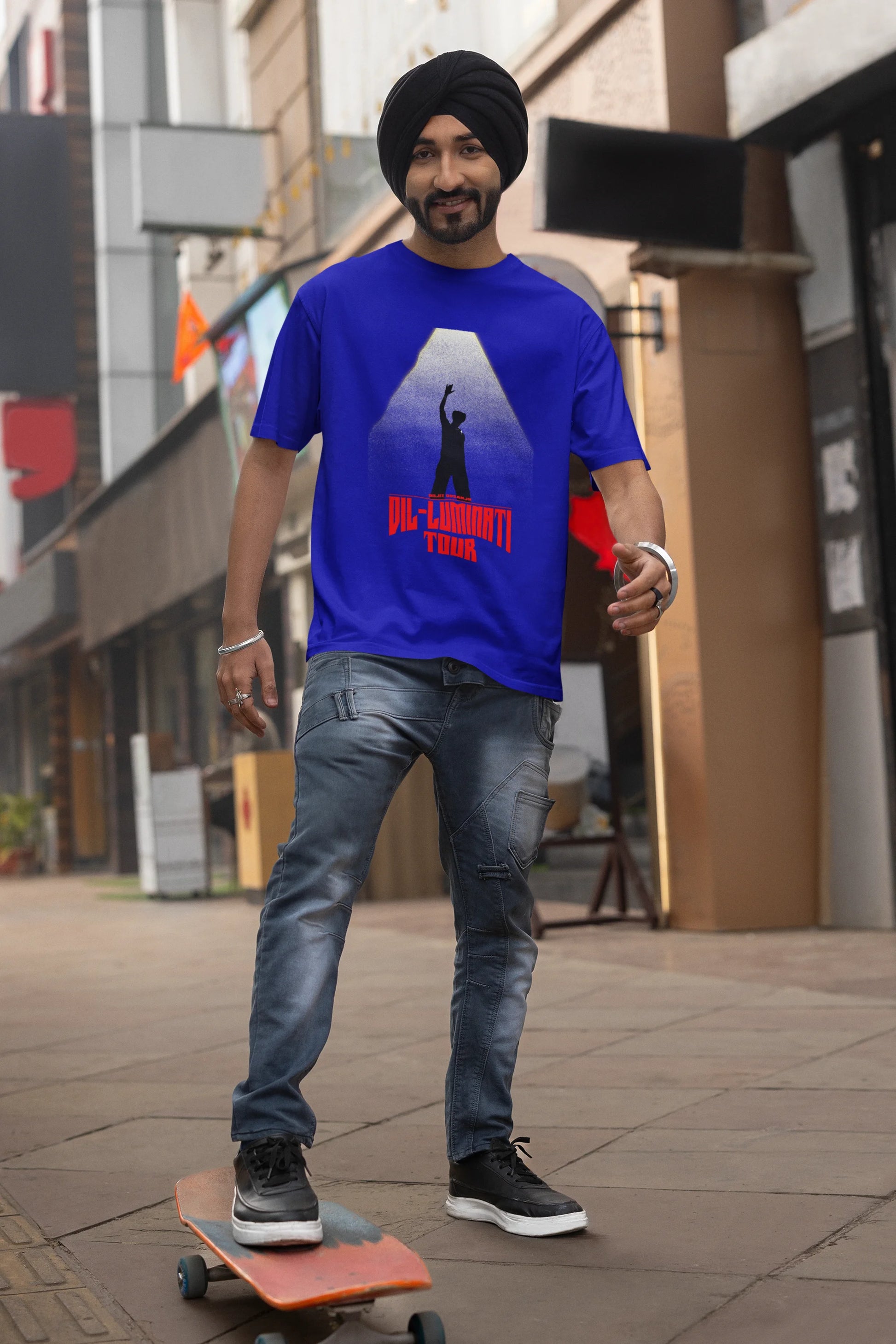 First front view of a male model wearing a royal blue oversized t-shirt with a portrait of Diljit Dosanjh on stage, bathed in the warm glow of a spotlight.