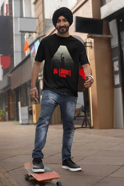 First front view of a male model wearing a black oversized t-shirt with a portrait of Diljit Dosanjh on stage, bathed in the warm glow of a spotlight.