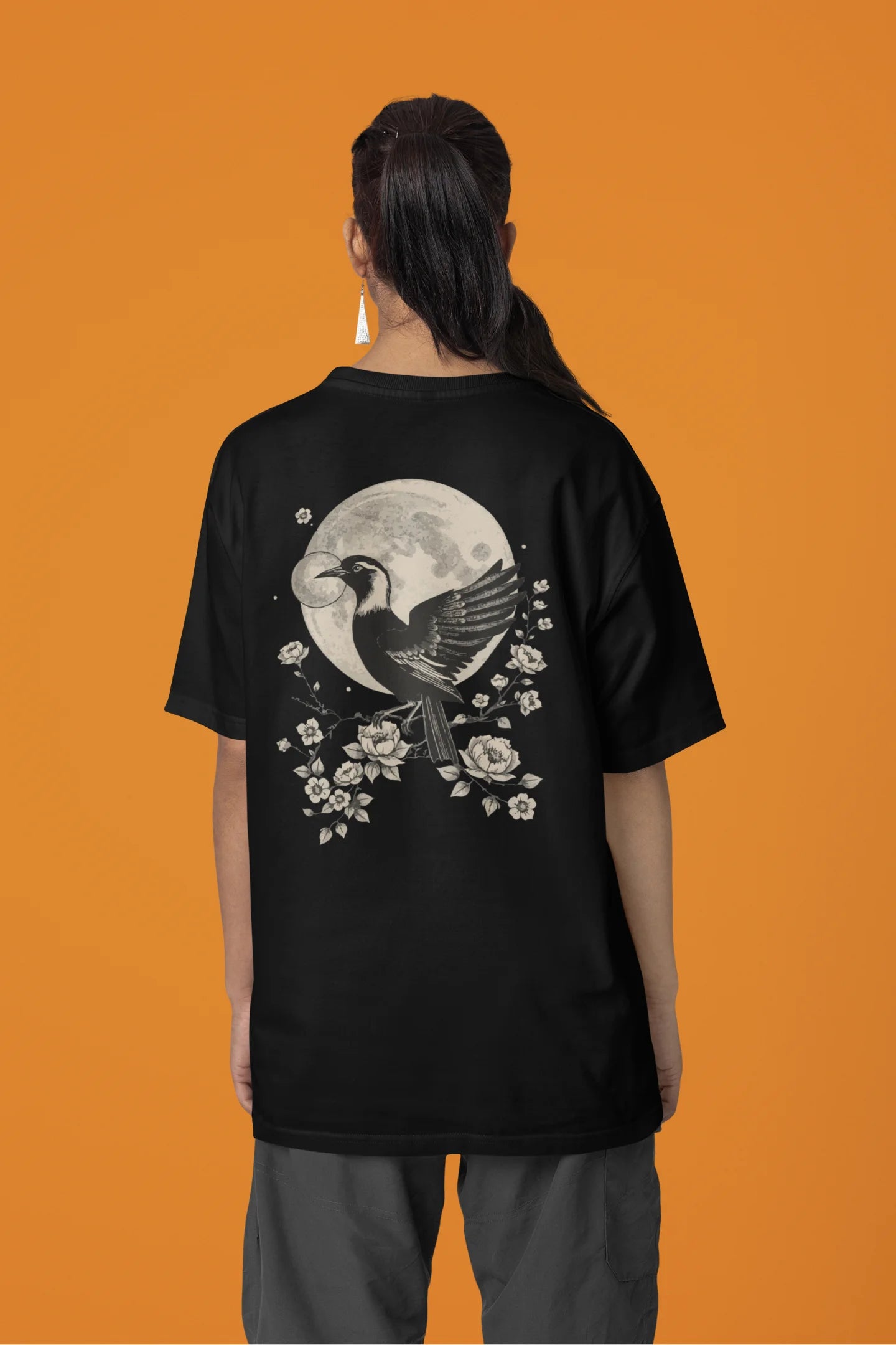 Back view of a female model wearing a black oversized tee with a heron taking off under the moonlight.