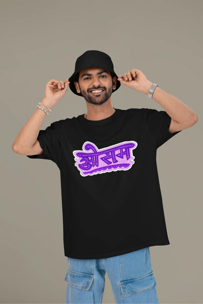 First front view of a male model wearing a black colored oversized graphic t-shirt featuring the word "awesome" in a Hindi script.