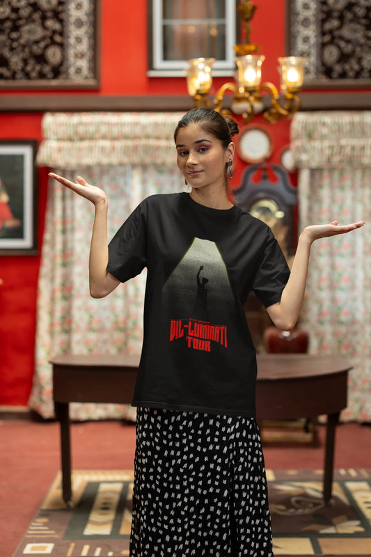 Second front view of a woman model wearing a black oversized t-shirt with a portrait of Diljit Dosanjh on stage, bathed in the warm glow of a spotlight.