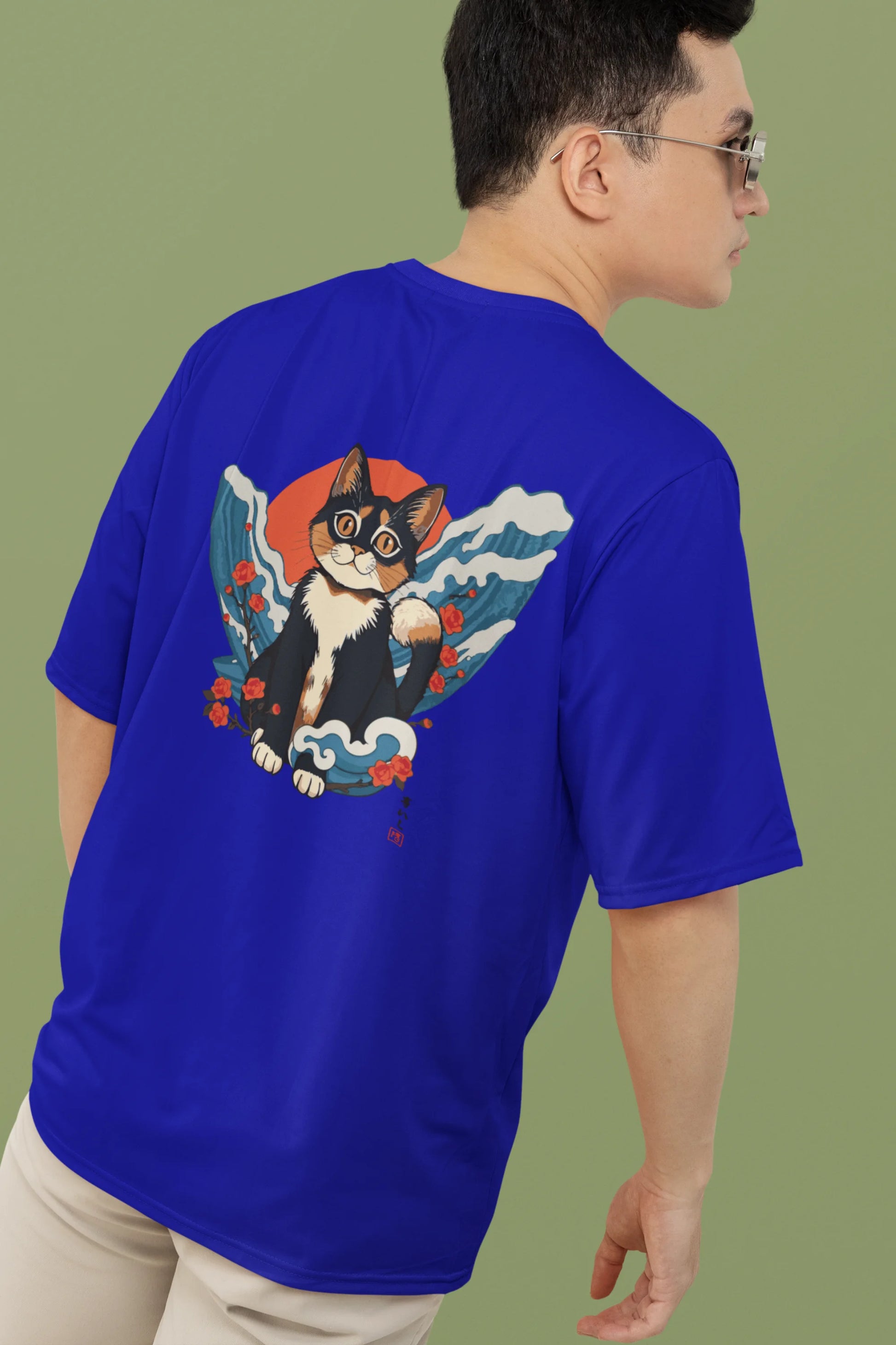 Back view of a male model wearing a royal blue oversized tee with a design of a cat dressed as a samurai, standing against a backdrop of waves and a rising sun.