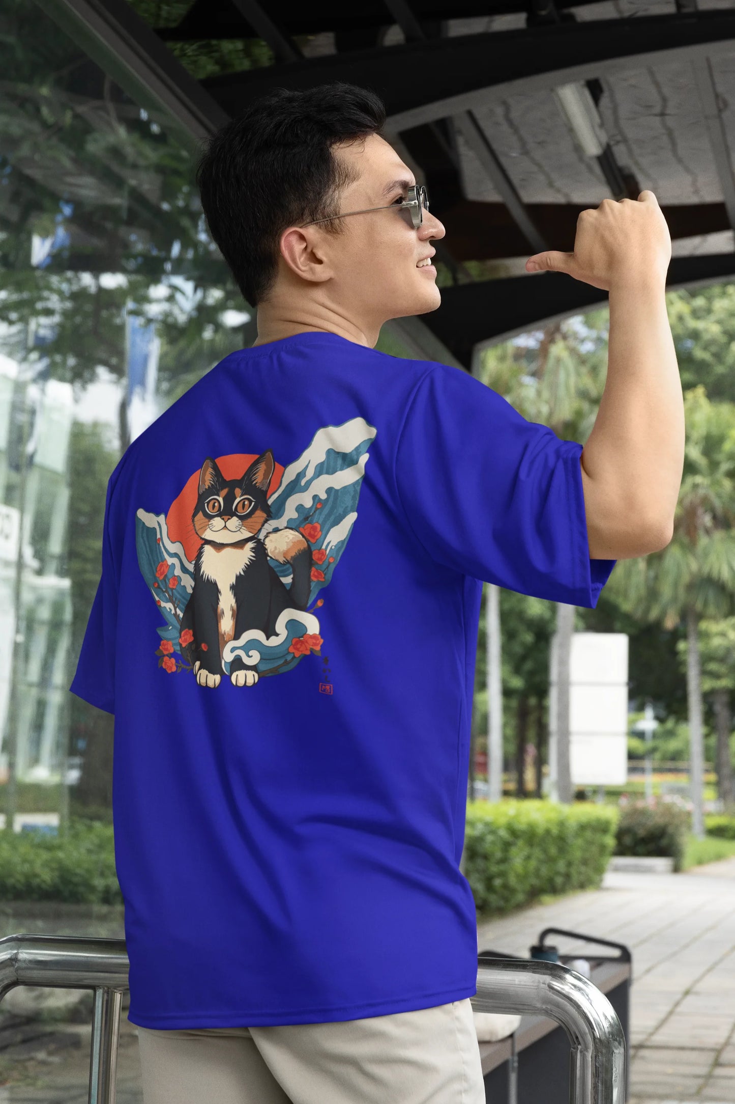 2nd back view of a male model wearing a royal blue oversized tee with a design of a cat dressed as a samurai, standing against a backdrop of waves and a rising sun.