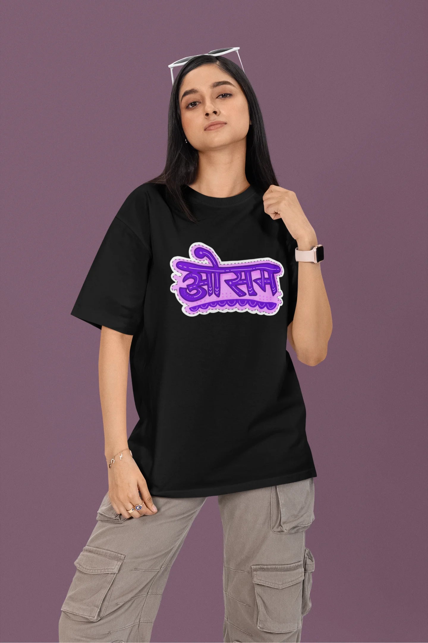 First front view of a female model wearing a black colored oversized graphic t-shirt featuring the word "awesome" in a Hindi script.